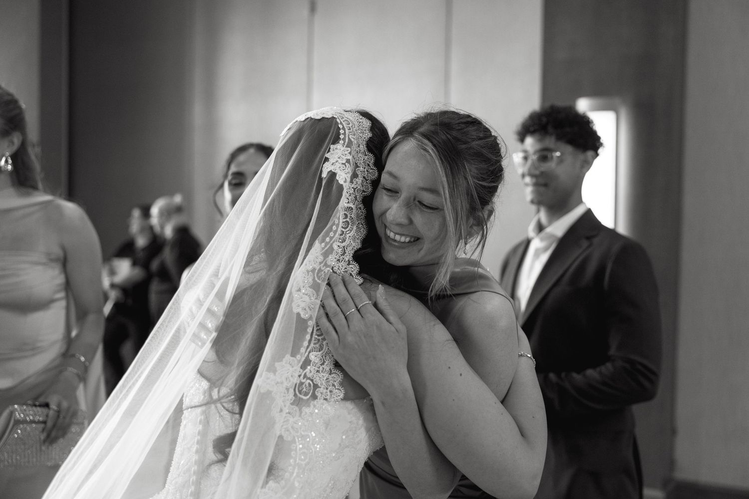 Bride in lace gown and veil shares emotional hug with guest at wedding ceremony in black and white photo.
