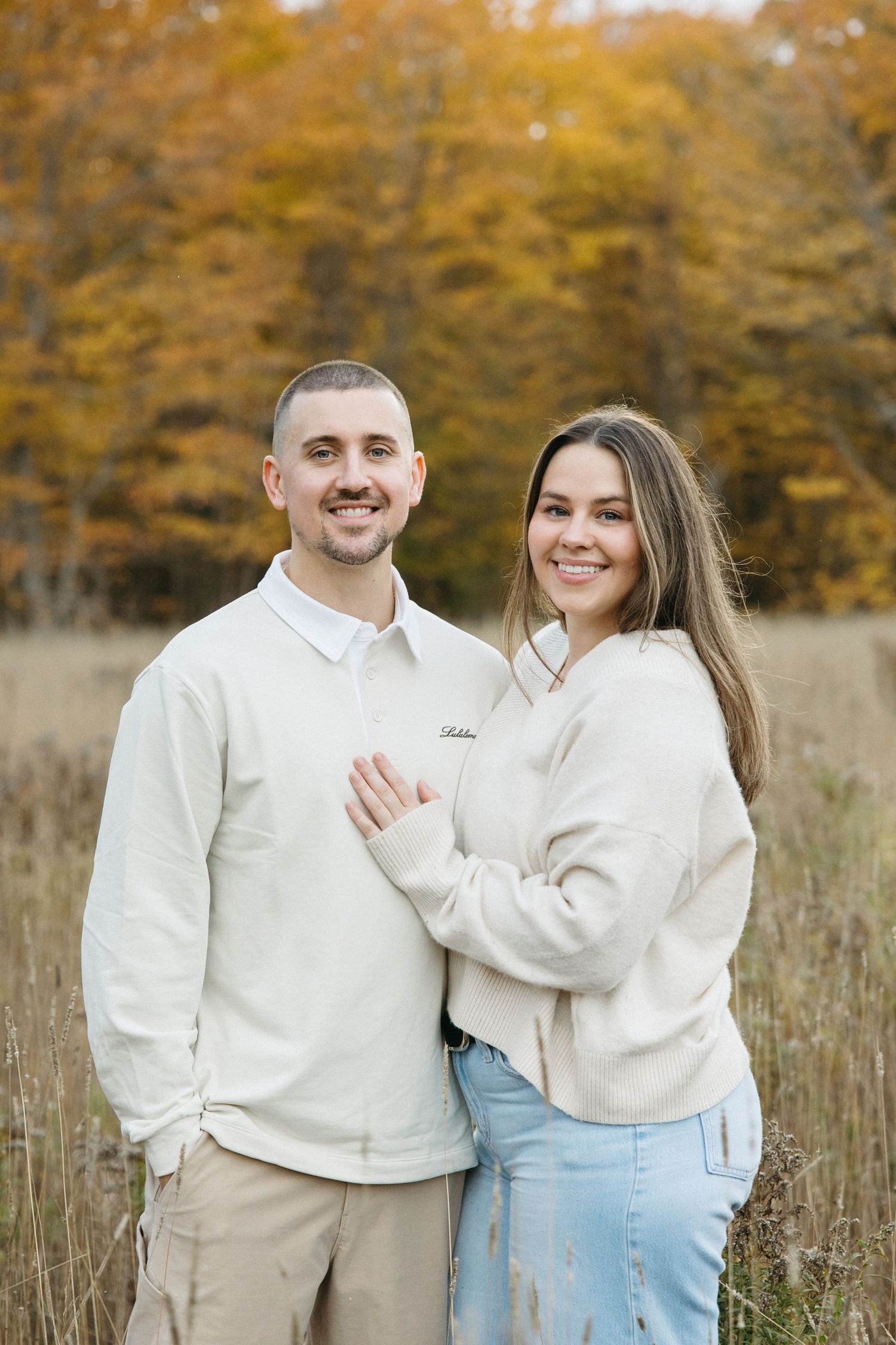A couple wearing coordinated white sweaters pose together in a golden autumn field.