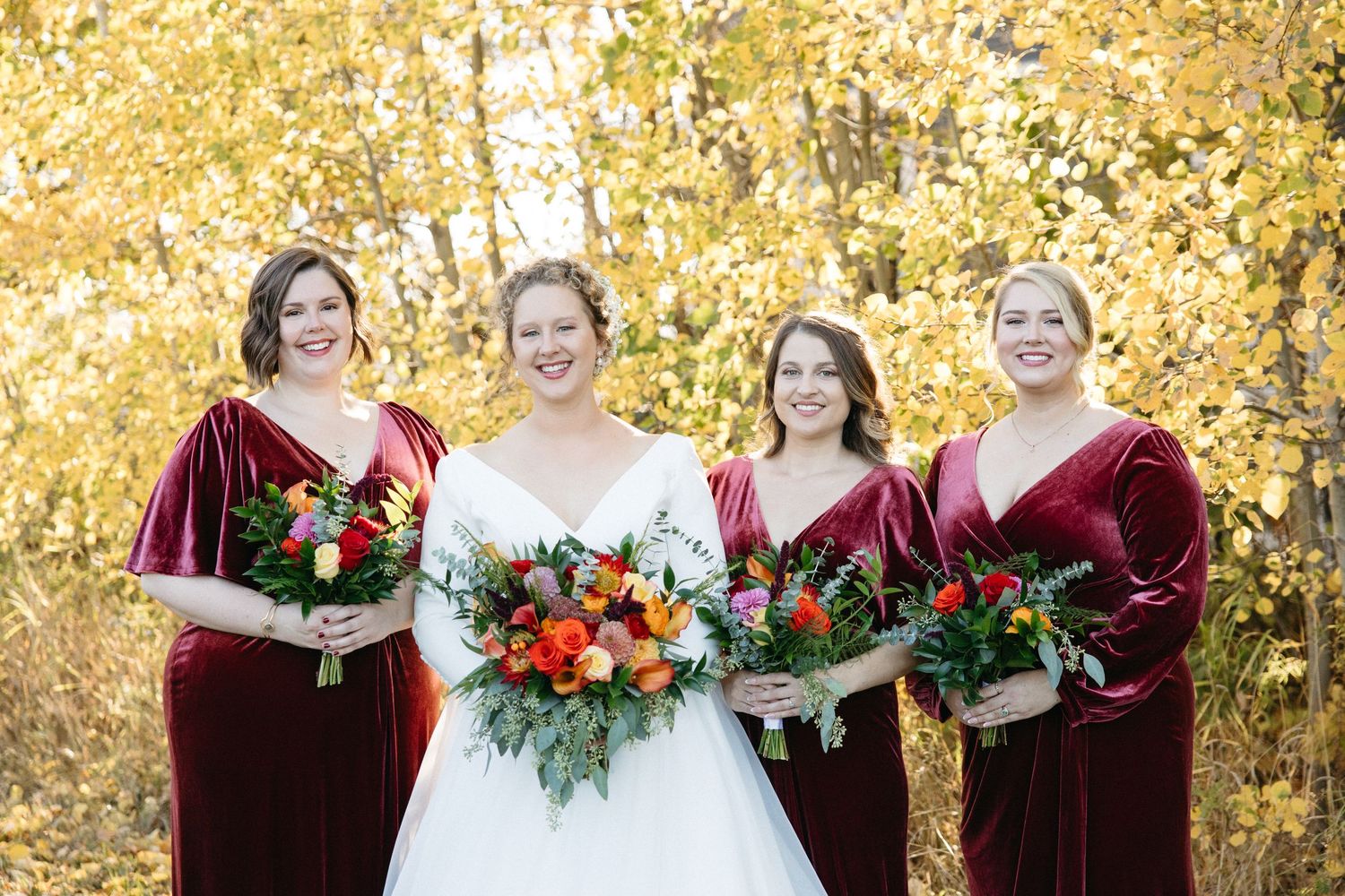 Wedding party in burgundy velvet dresses poses against golden autumn foliage.