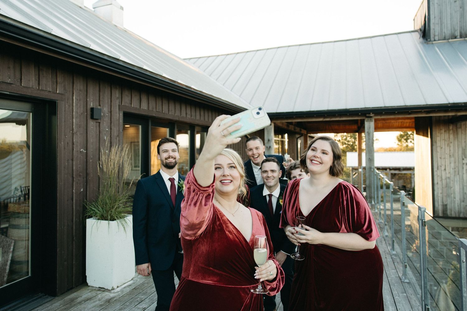 Wedding party in burgundy dresses and dark suits poses outside a rustic wooden venue.