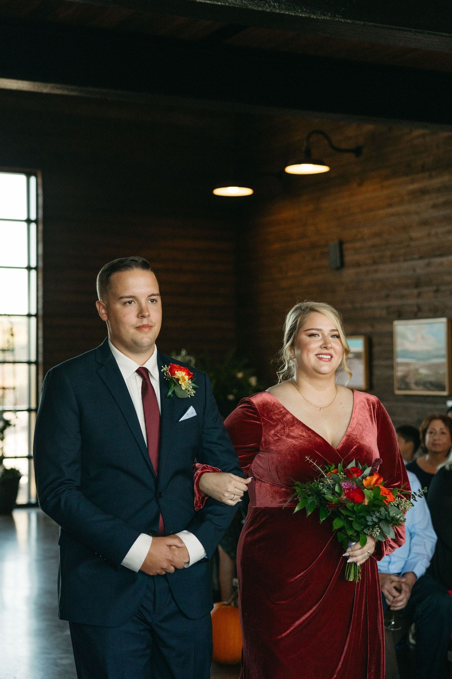 Wedding attendees in formal attire gather in a brick-walled venue with natural lighting.