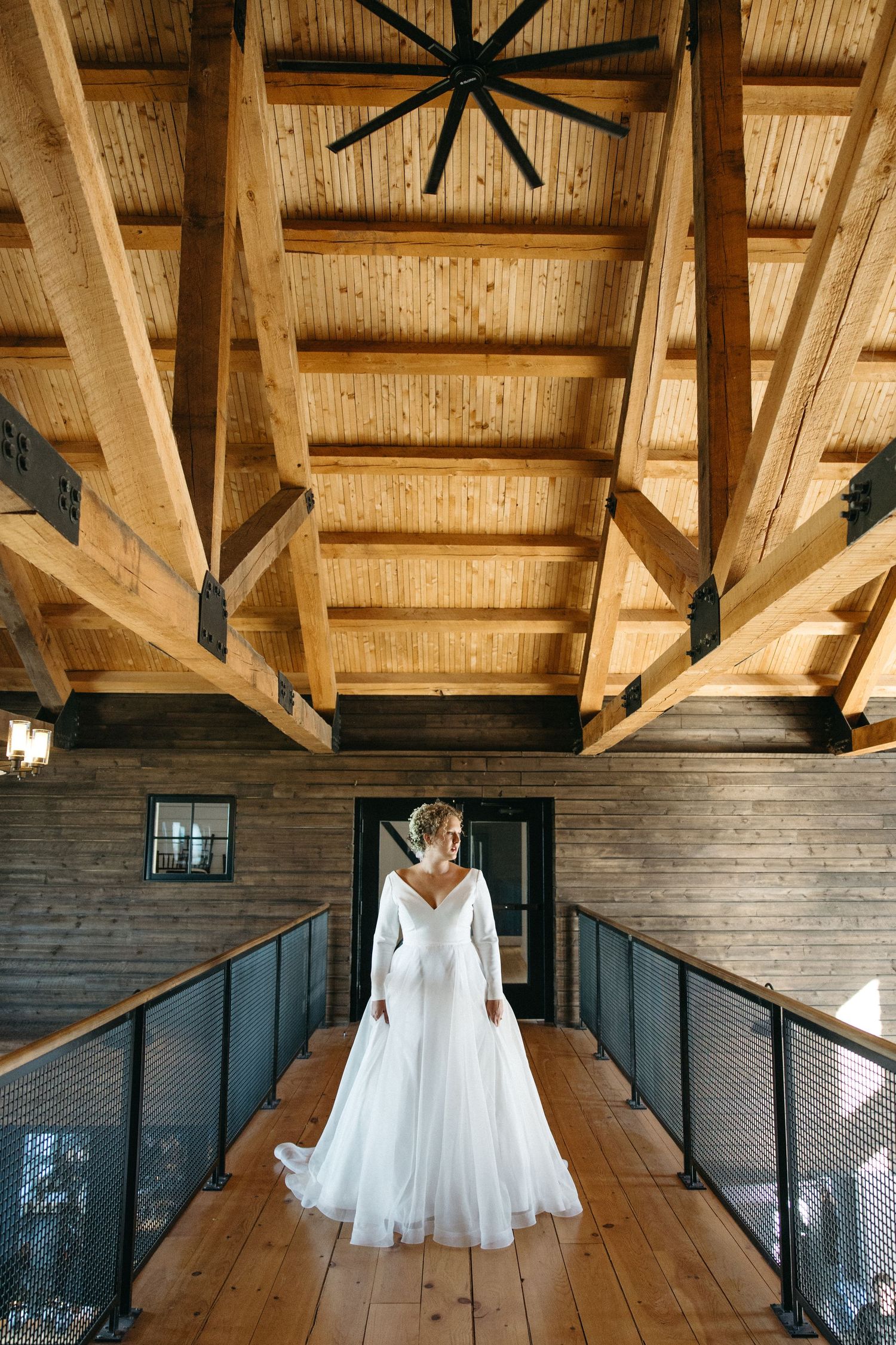 A bride in a flowing white gown stands on a wooden walkway under rustic wooden beams.
