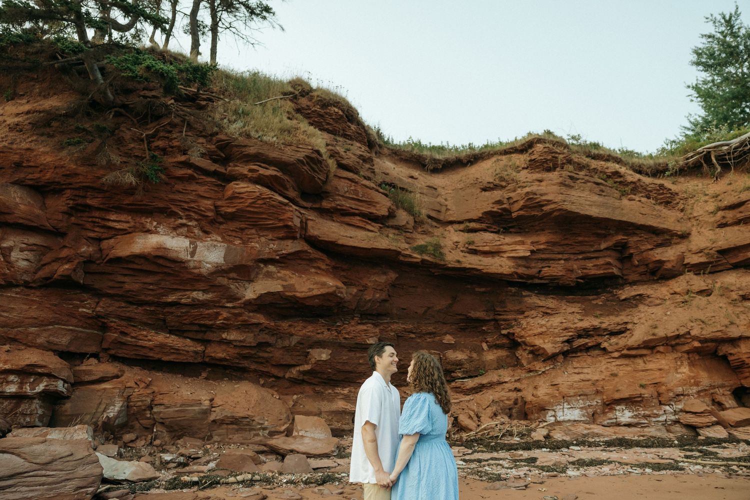 A couple walks together along a scenic beach with red rocky cliffs in the background.