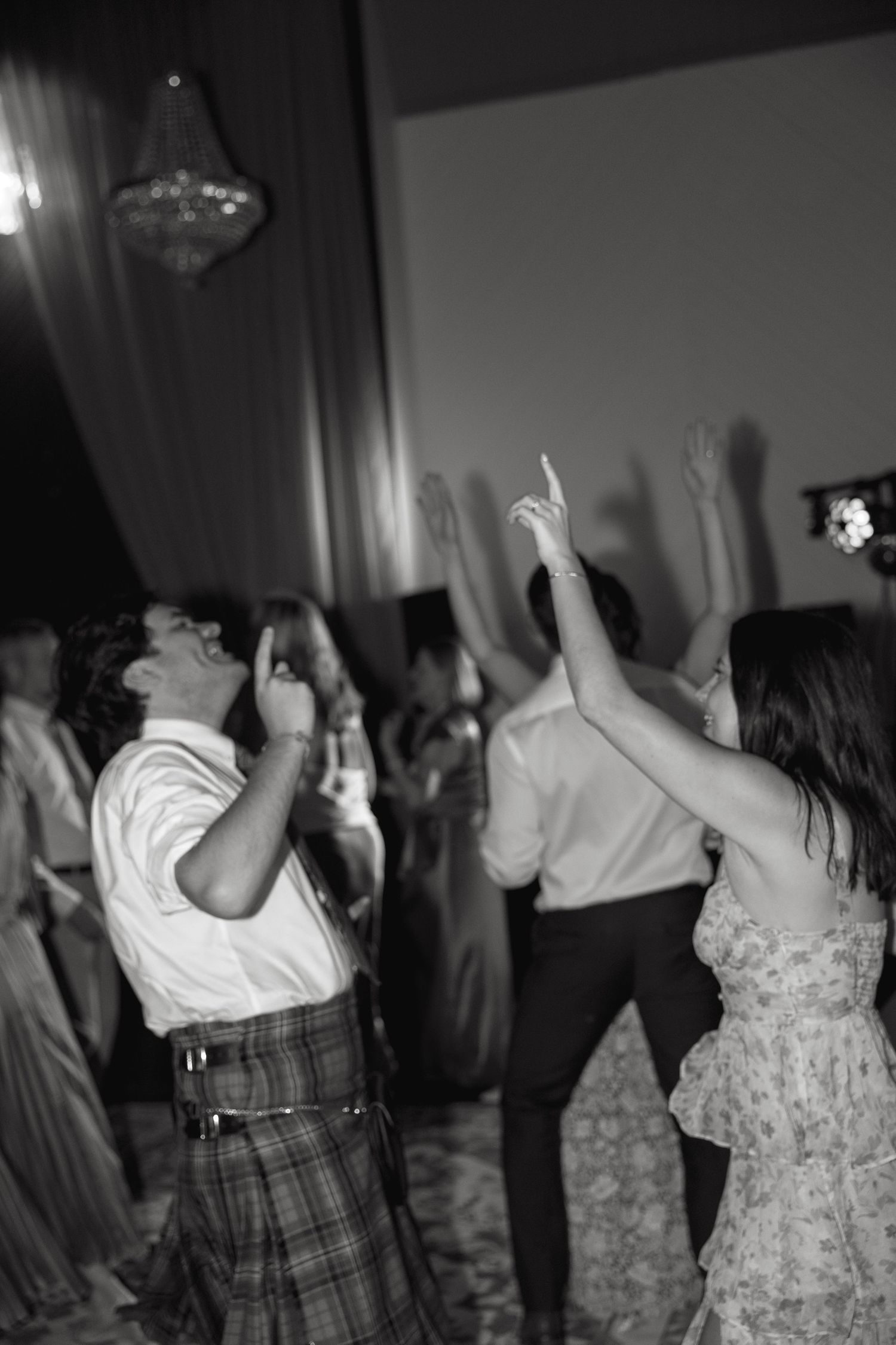 Wedding guests dancing with raised hands under a crystal chandelier in black and white.