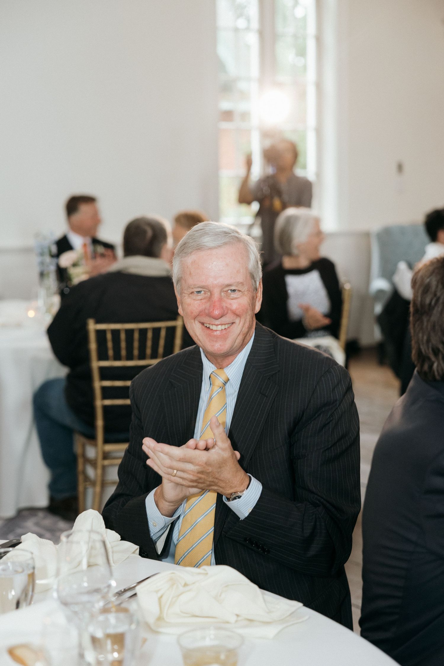 A wedding reception guest in formal attire smiling and clapping at a table.