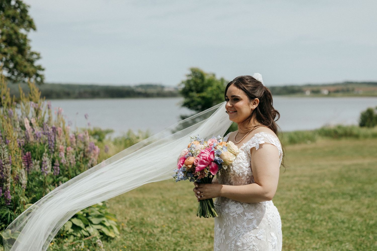 A bride in a white dress holds a pink bouquet against a coastal landscape.