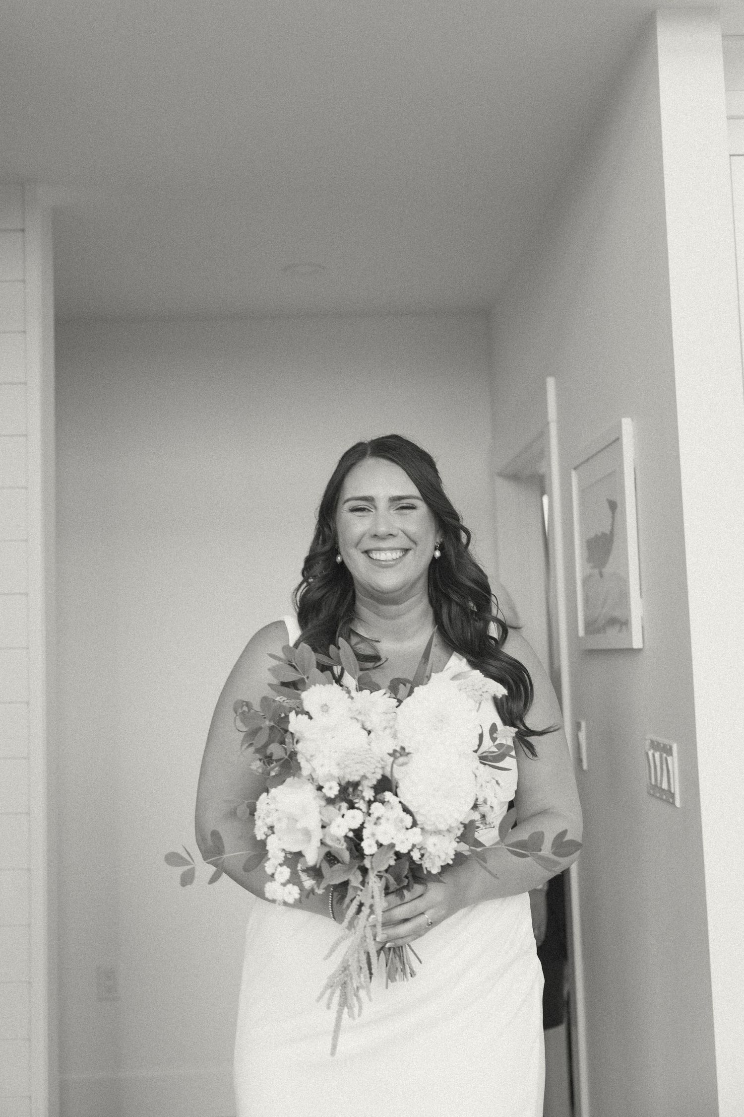A bride holds a white floral bouquet while standing in a bright modern interior space.
