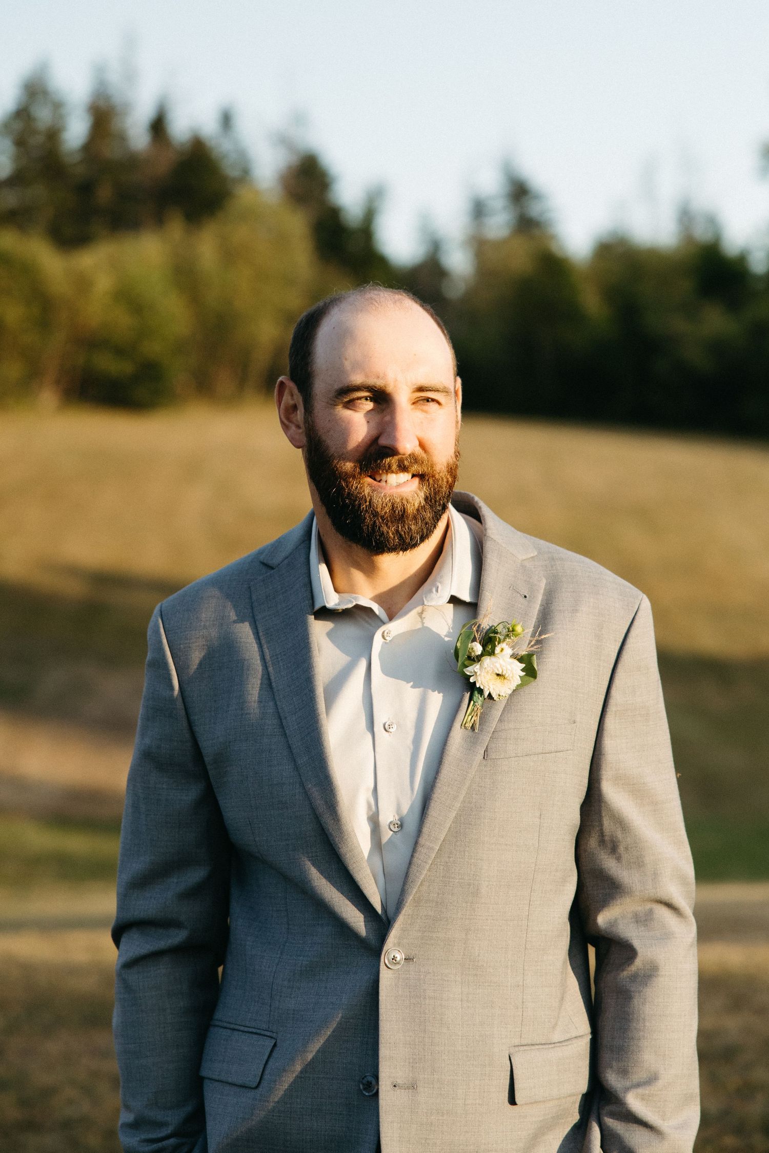 Portrait of a groom in a light grey suit with boutonniere smiling in golden sunlight.