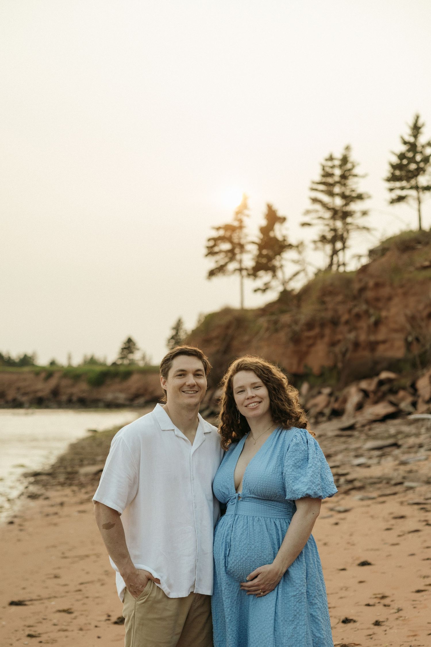 A couple poses on a beach at sunset with pine trees and red cliffs in the background.