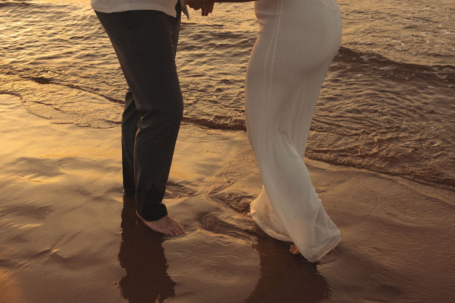 Couple holding hands walking barefoot on a beach at sunset.