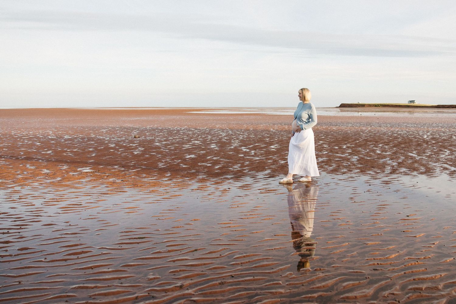 Woman in white dress and blue jacket stands on reflective wet sand beach under a cloudy sky.