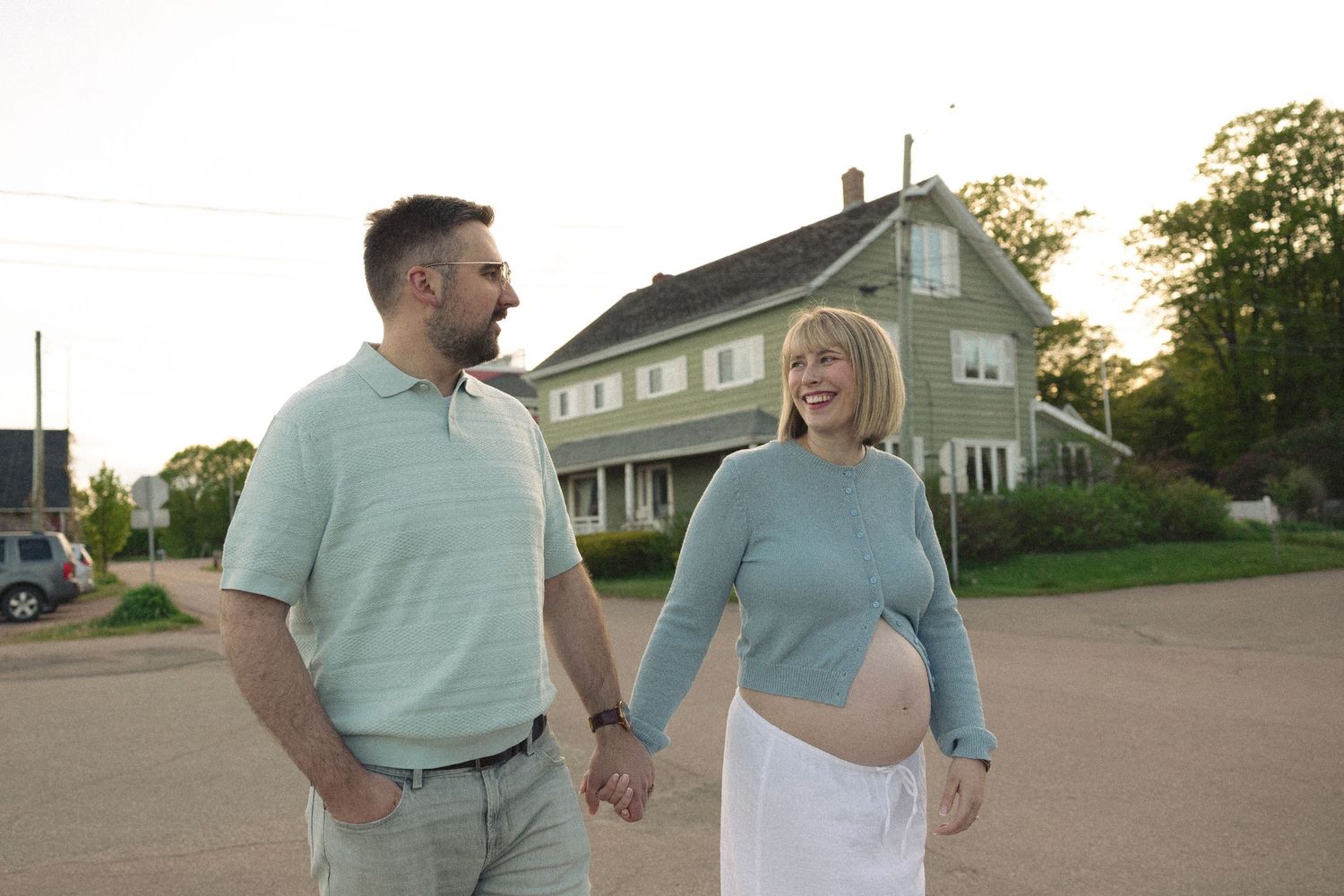 Pregnant couple holding hands and smiling outdoors near a house at sunset.