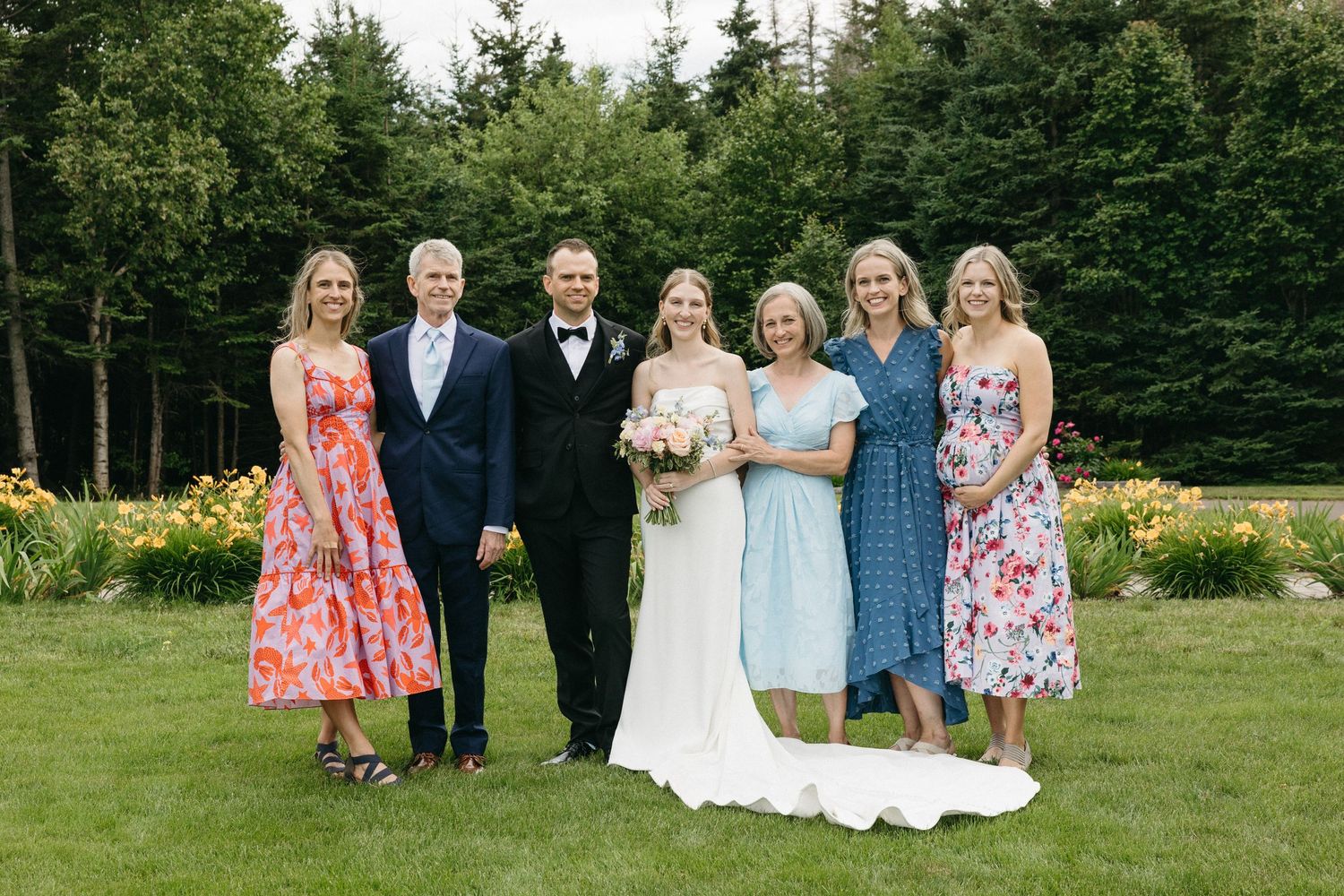 Wedding party of six poses together in garden with colorful floral dresses and blooming flowers.