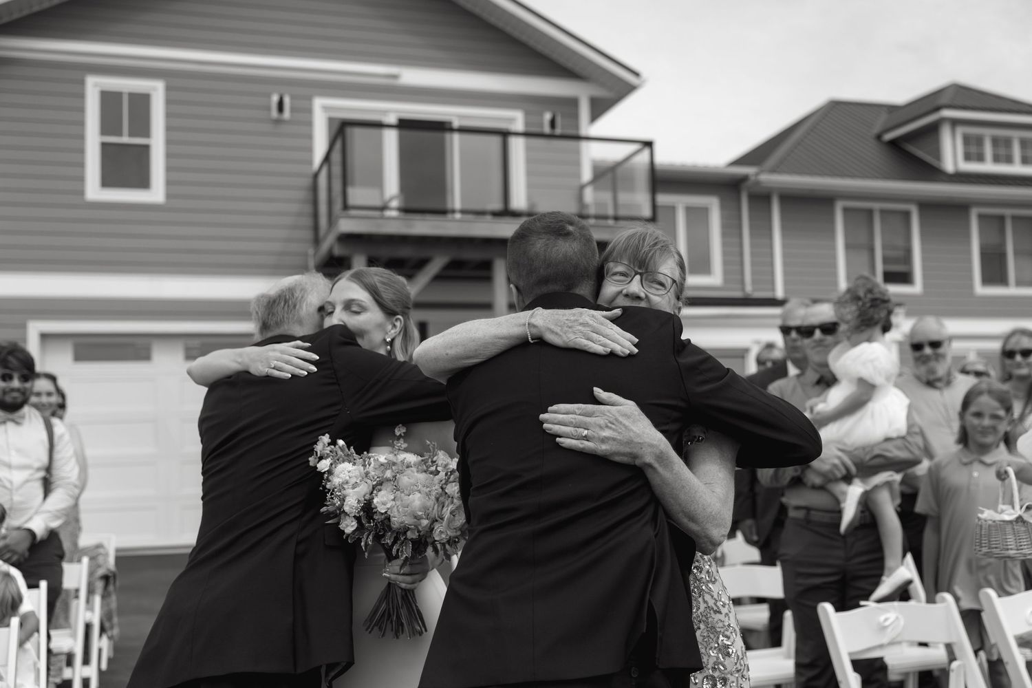 Groom sharing emotional group hug with family members during outdoor wedding ceremony in black and white.