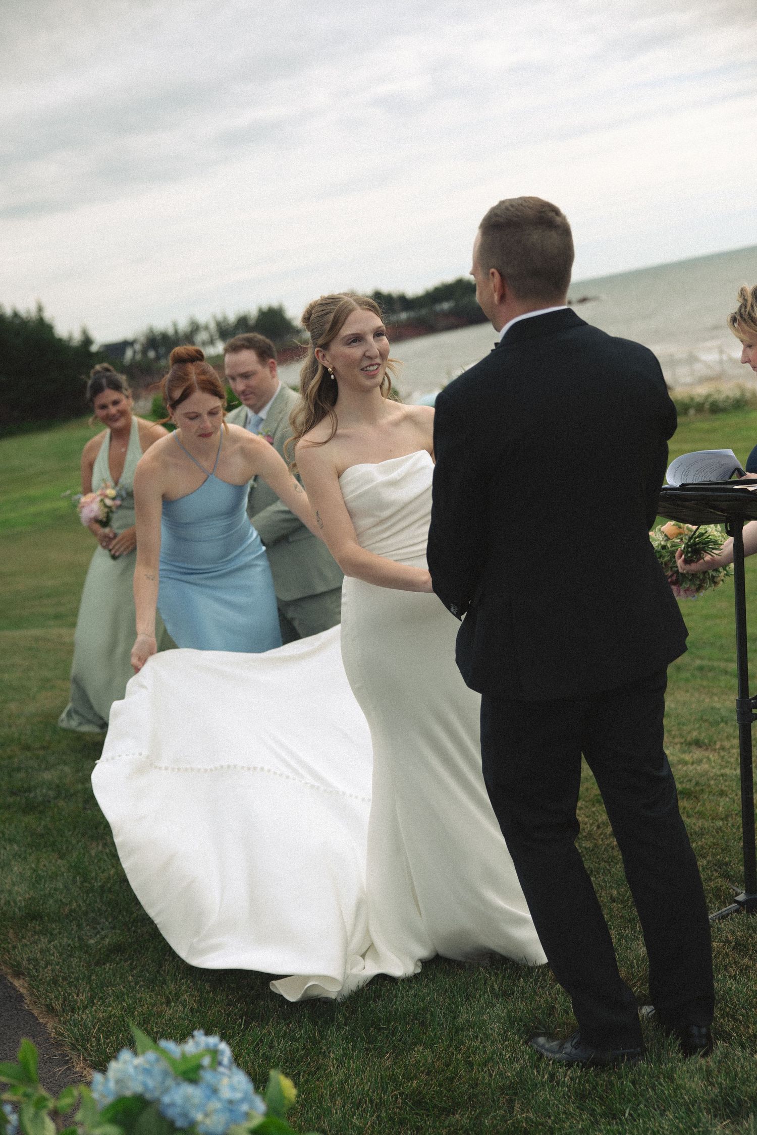 Smiling bride in strapless white gown approaches groom at outdoor wedding ceremony by the sea.