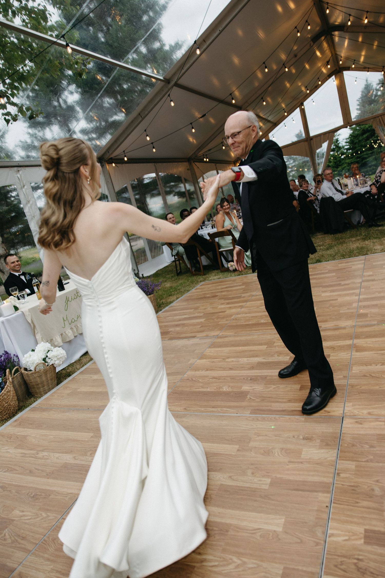 Bride in fitted white gown dancing with bald man in black suit on wooden dance floor under tent.