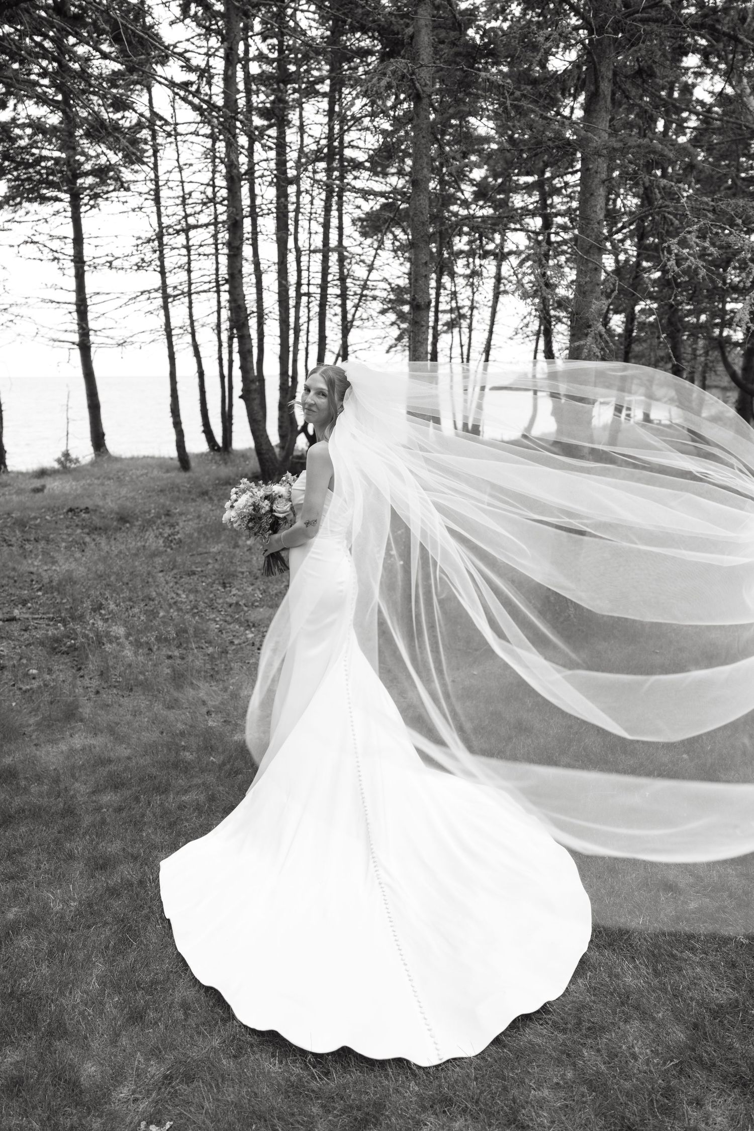 Black and white photo of bride in fitted gown with dramatic flowing veil among tall trees.
