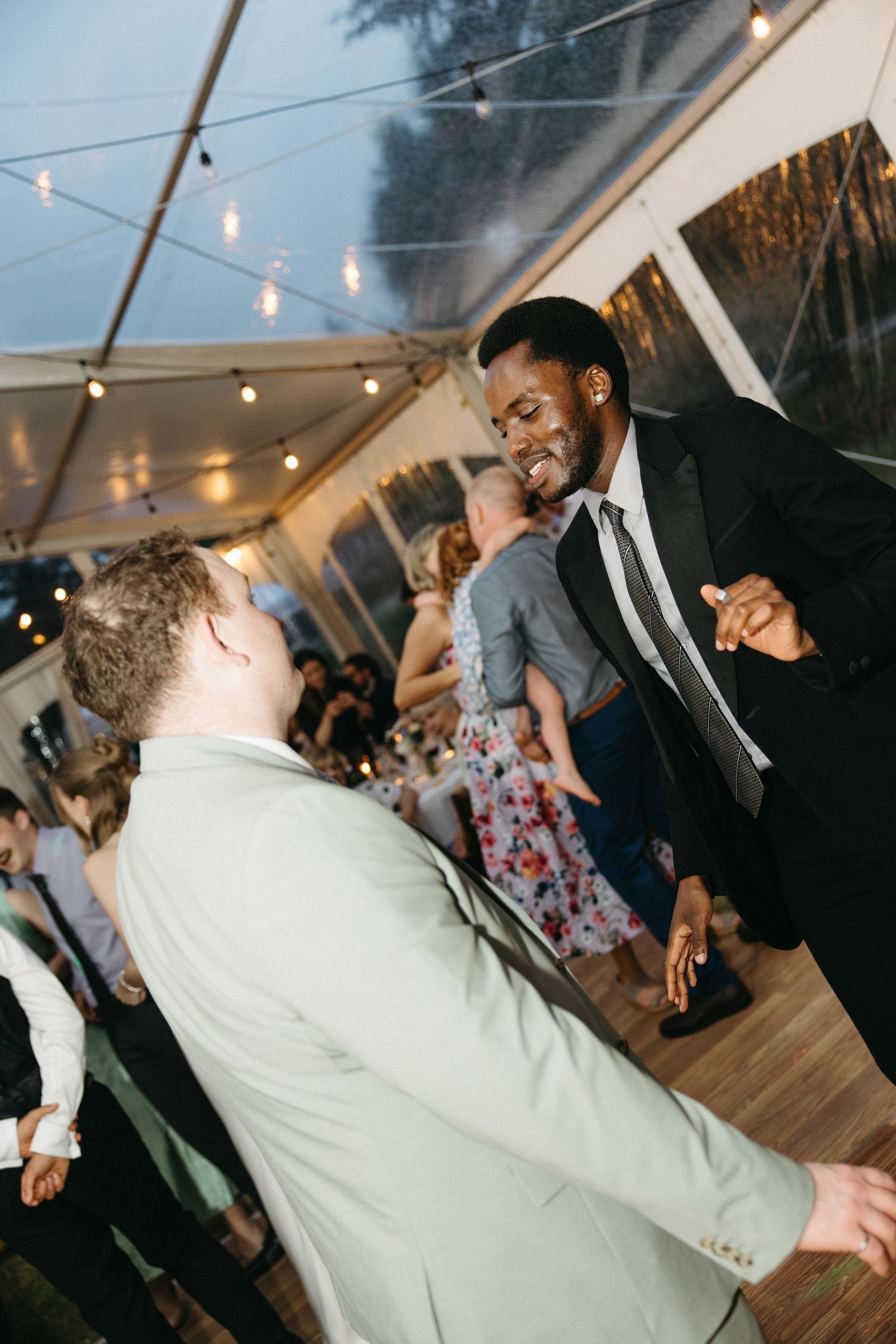 Two men dancing energetically at a wedding reception under string lights in a tent.