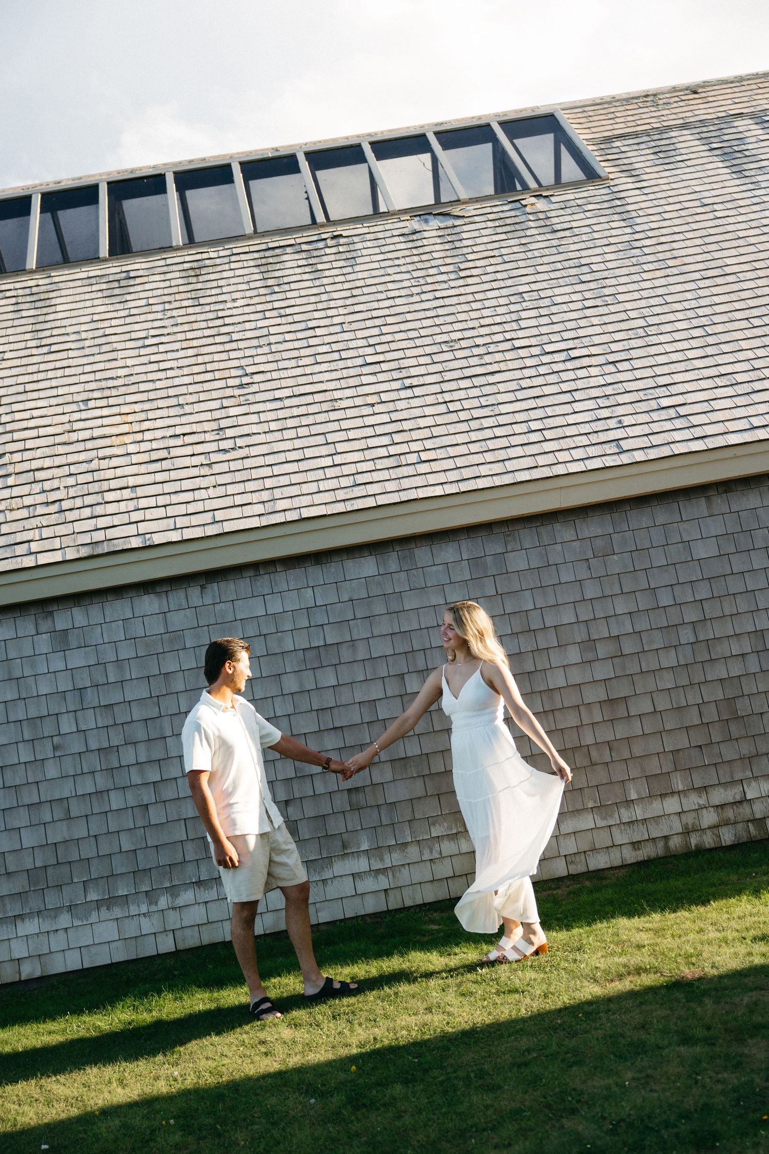 Couple holding hands and walking on green grass beside a shingled building on a sunny day.