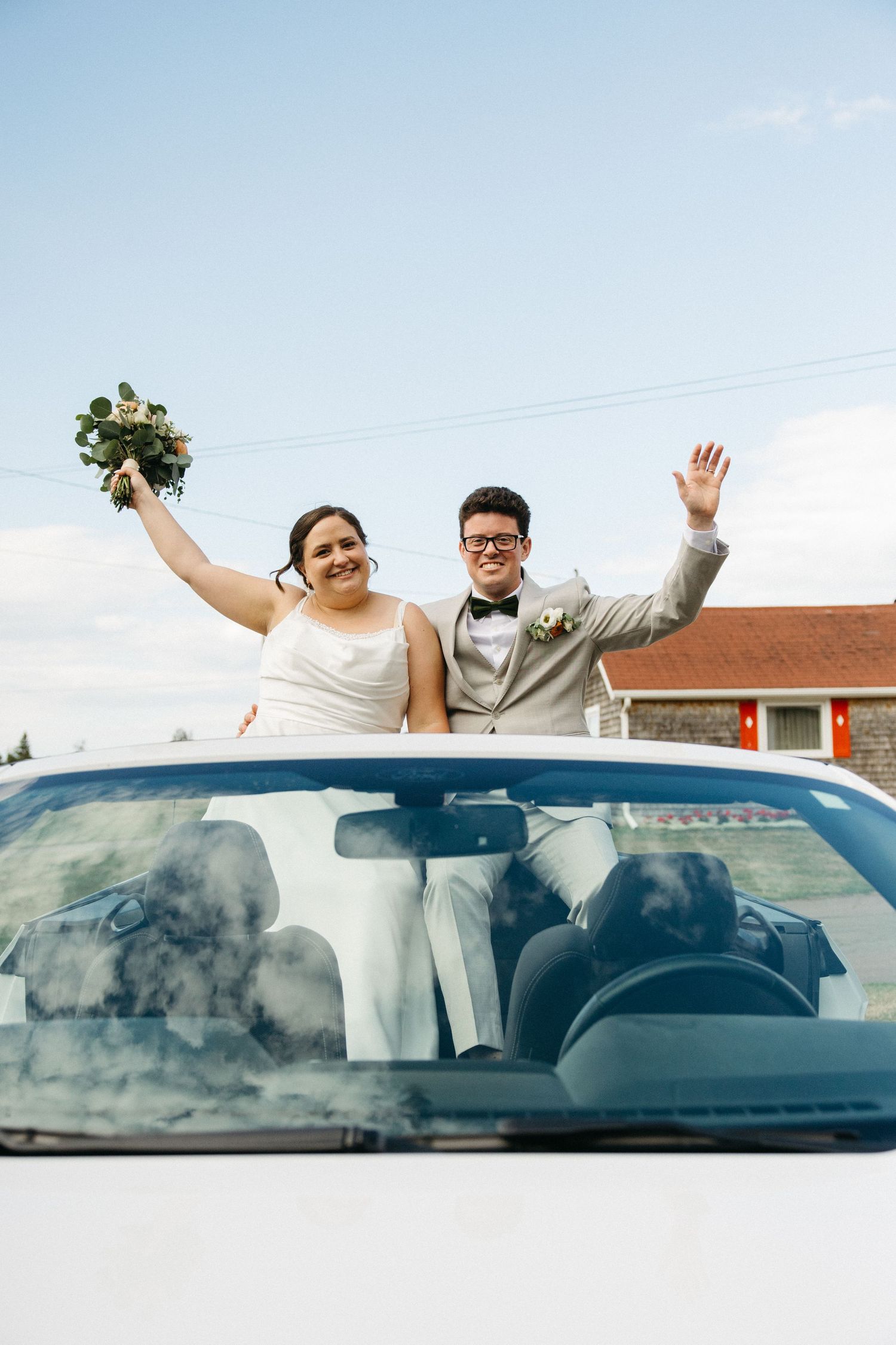 Happy bride and groom wave from white convertible car after their outdoor wedding ceremony.