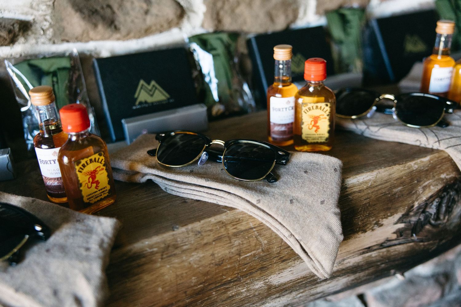 Groomsmen gifts displayed on rustic wood including Fireball whiskey bottles, sunglasses, and green pouches.
