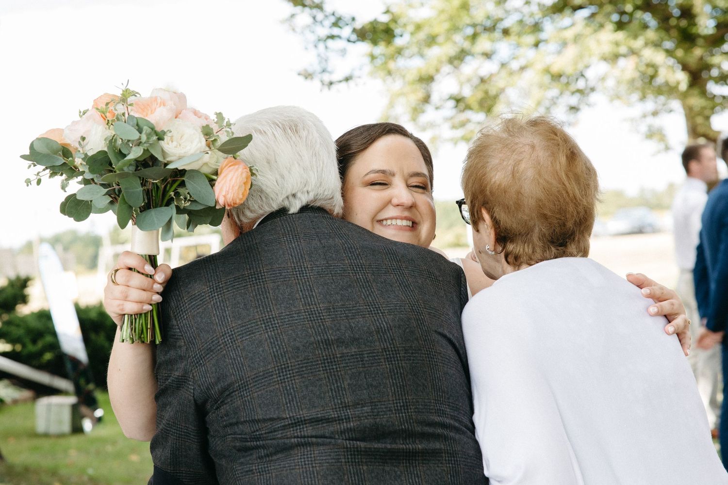 Smiling bride holding a peach bouquet embracing two elderly guests warmly at an outdoor wedding ceremony.