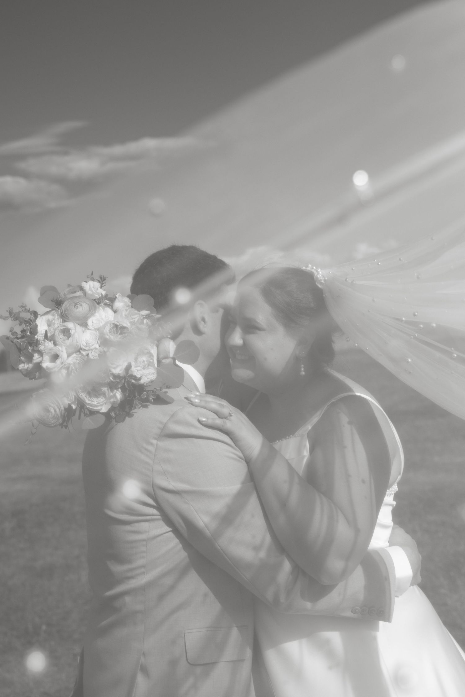 Dreamy black and white photo of bride and groom embracing under flowing veil outdoors.