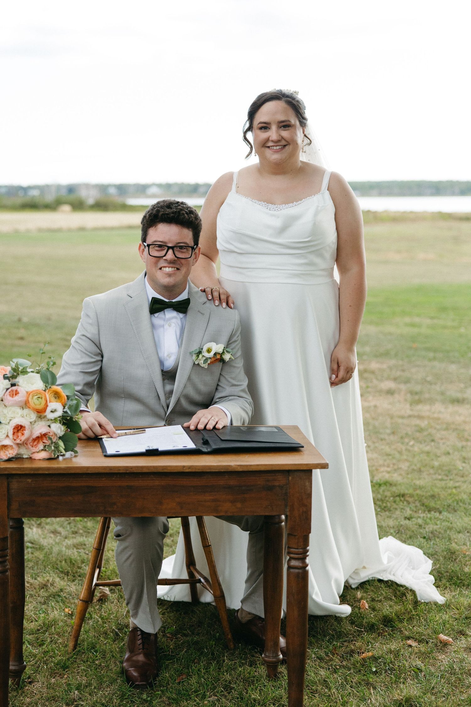Bride in white gown and groom in gray suit sign wedding documents at a wooden table outdoors.