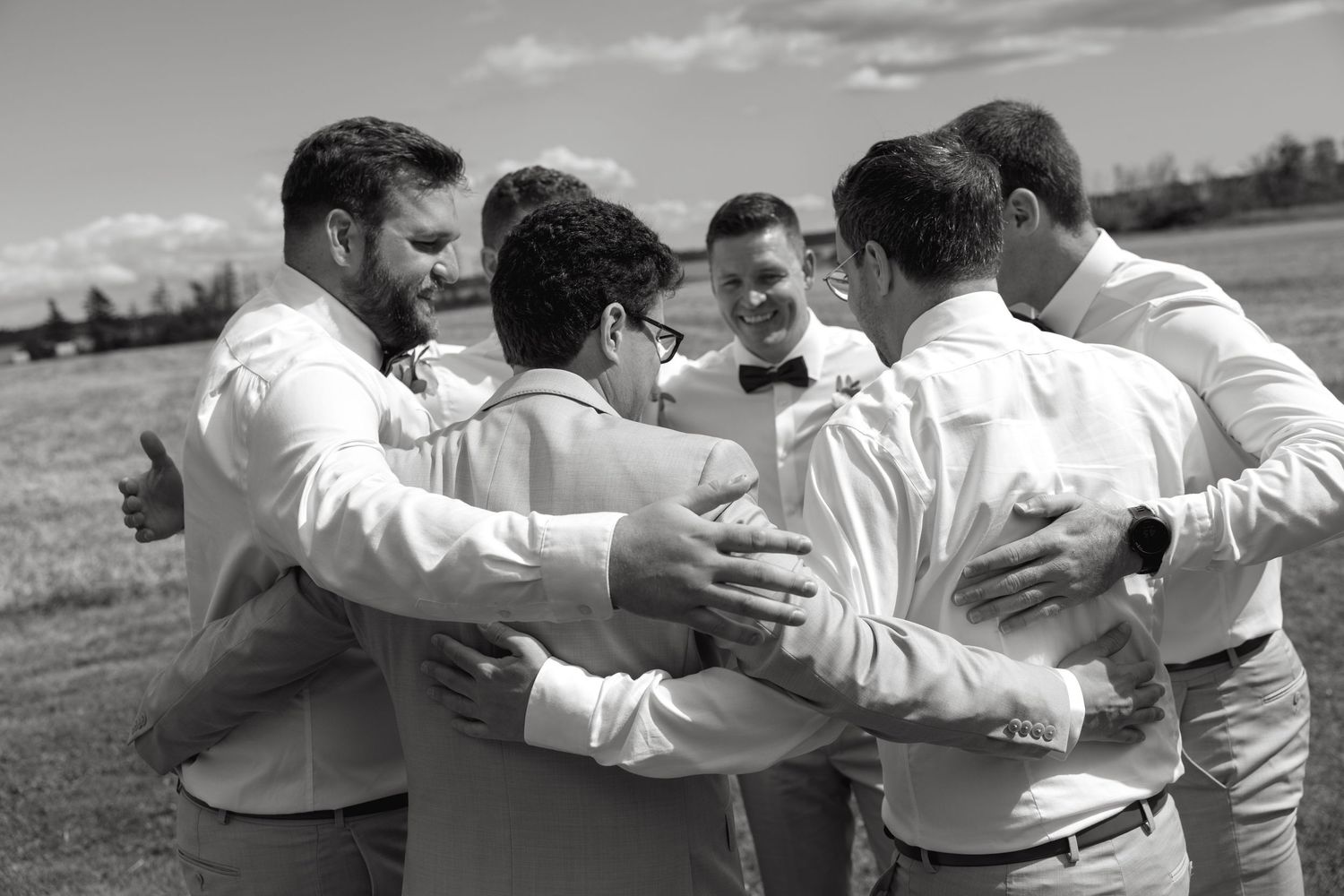 Black and white photo of groom and groomsmen sharing a group hug outdoors on wedding day.