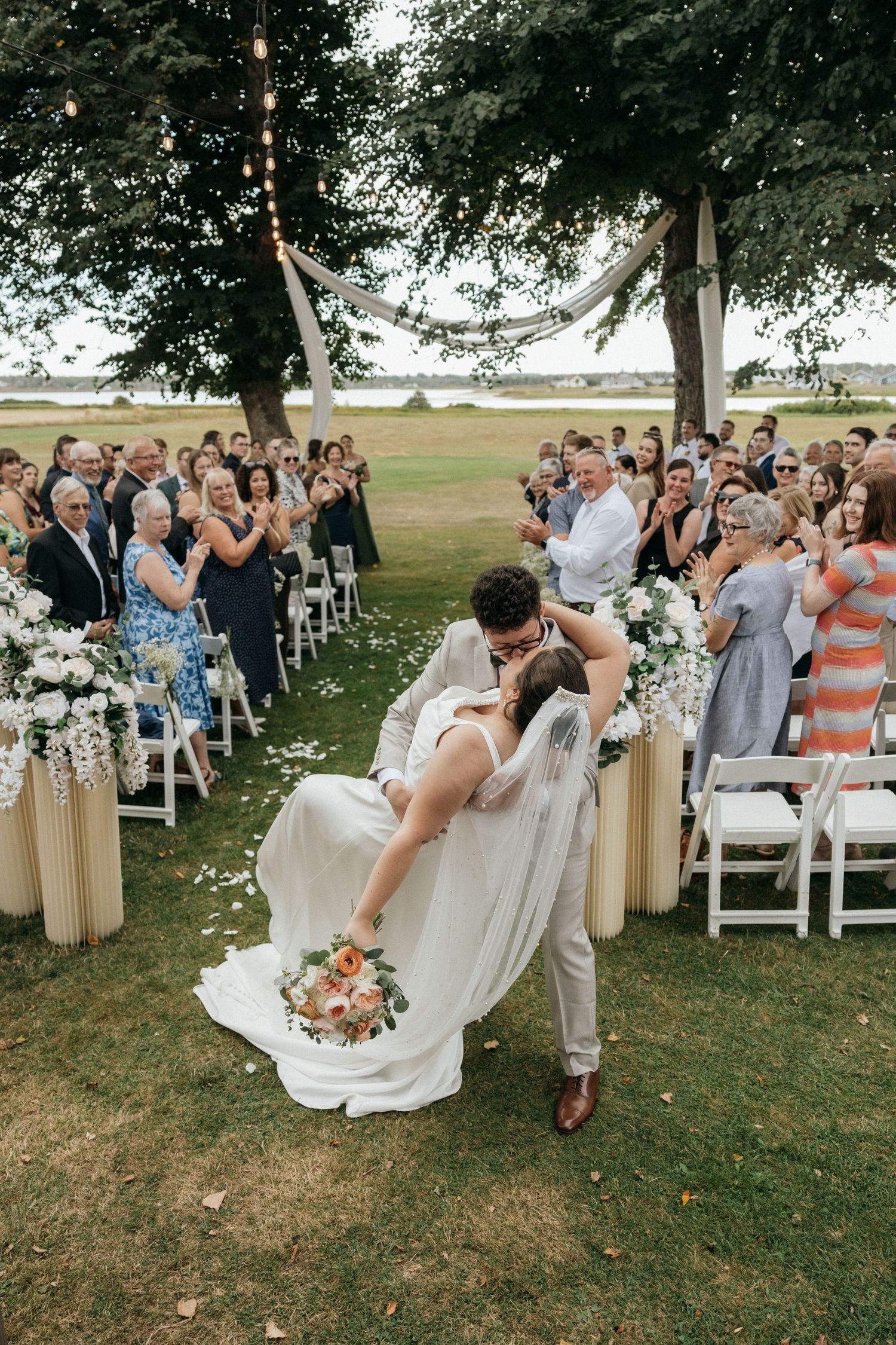 Bride and groom share a romantic dip kiss at outdoor wedding ceremony while guests cheer and applaud.