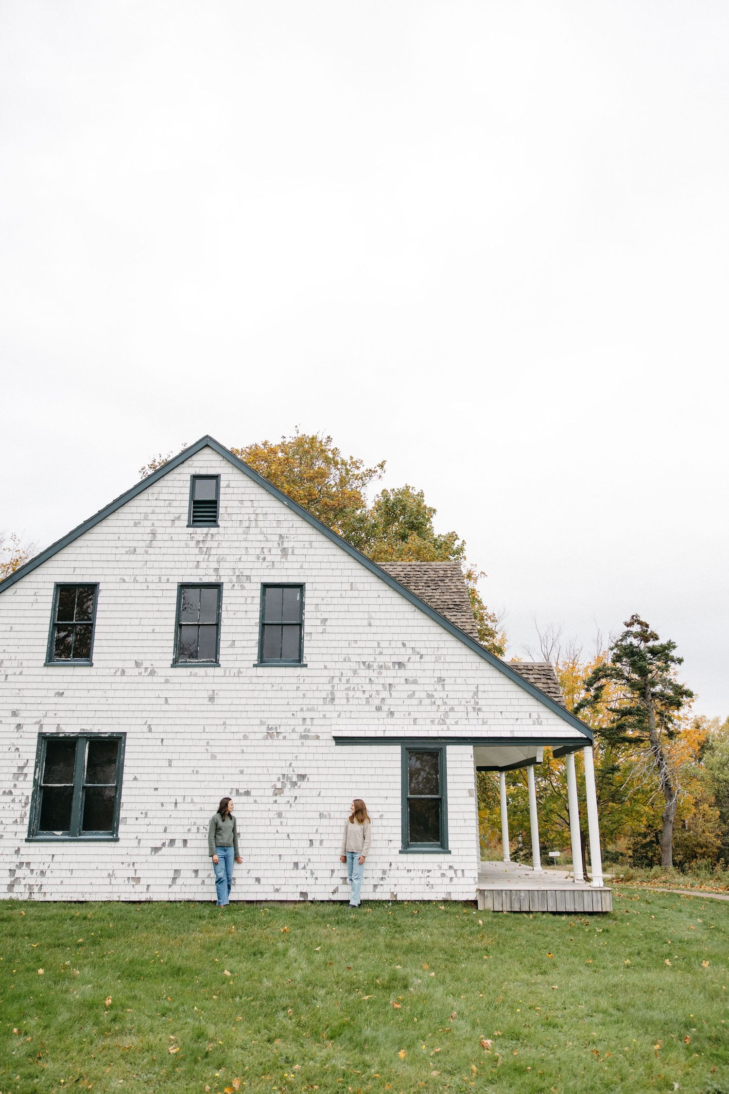 Two people in jeans stand against a white shingled farmhouse with green trim and a porch, surrounded by autumn trees and a lawn.