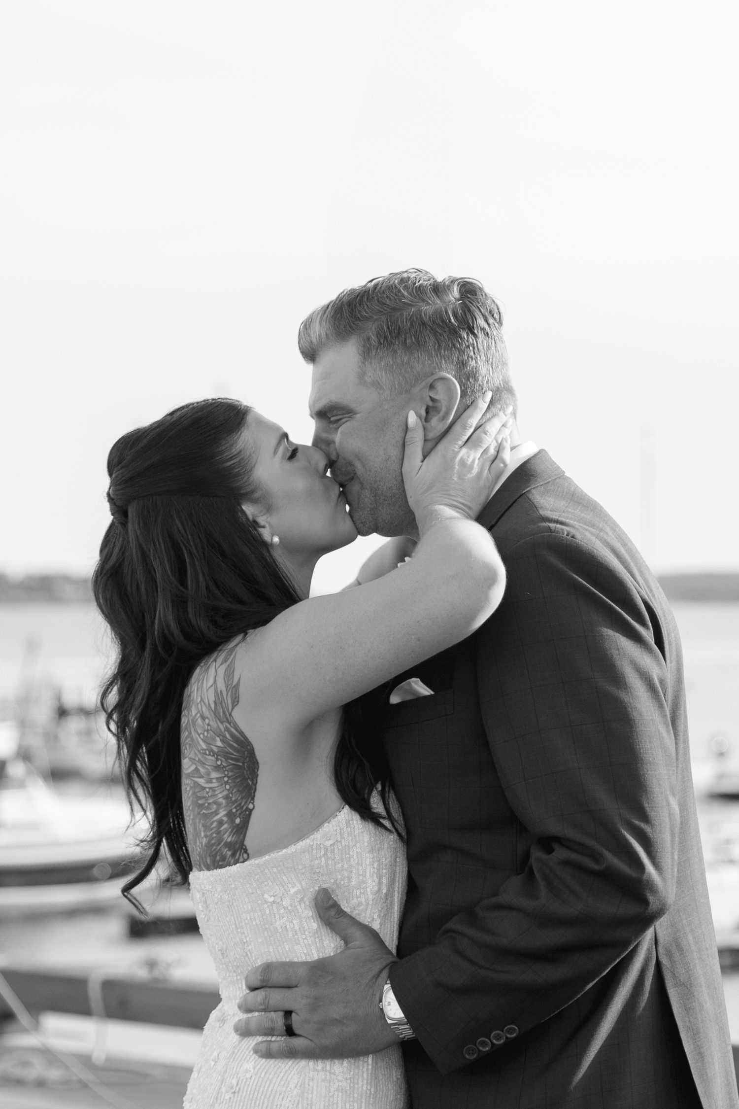 Black and white photo of a bride in a lace gown and a groom in a suit sharing a kiss at a marina on their wedding day.