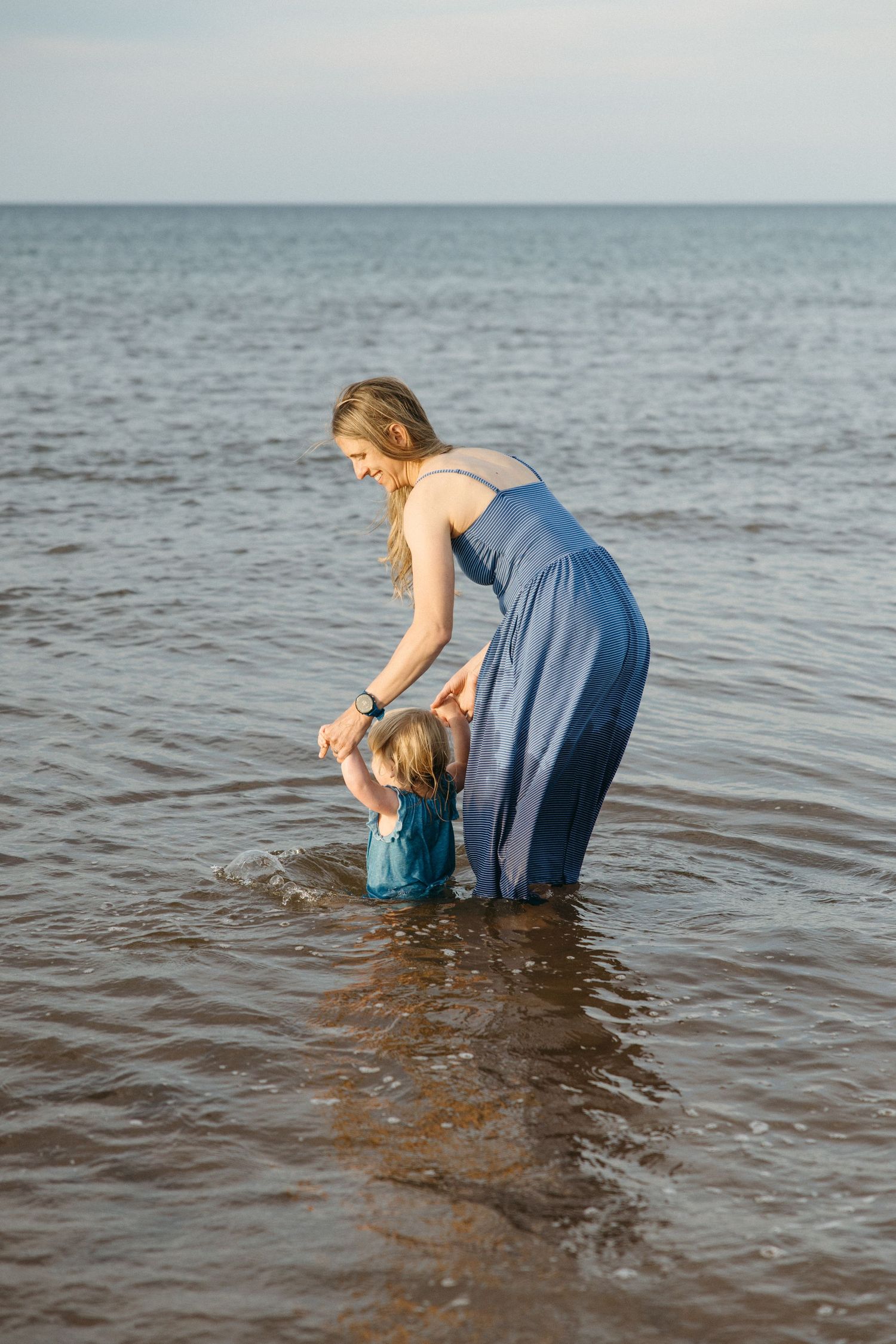 A smiling mother in a blue dress helps her toddler splash in shallow ocean waves at the beach.