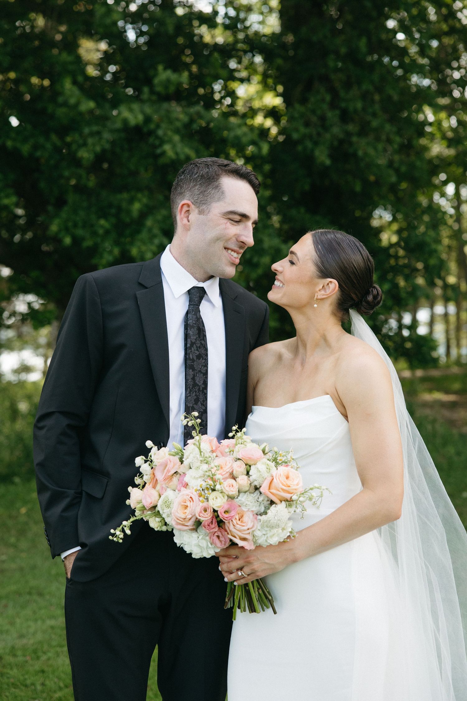 A smiling bride and groom share a romantic moment outdoors, she holds a pink and white bouquet, wearing a strapless gown and veil.