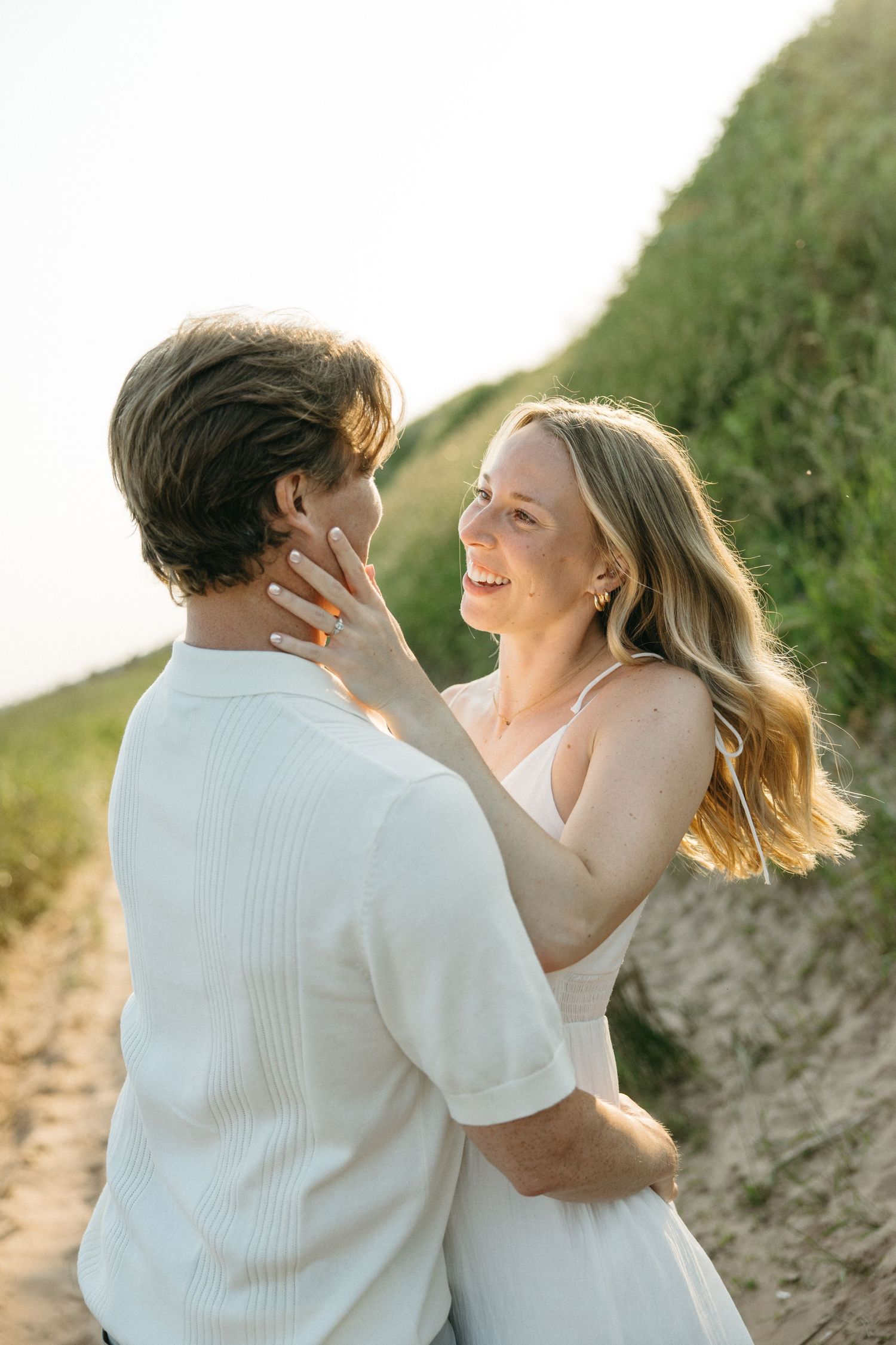 Couple embracing on a sandy beach path at golden hour, woman in white dress smiling up at man.