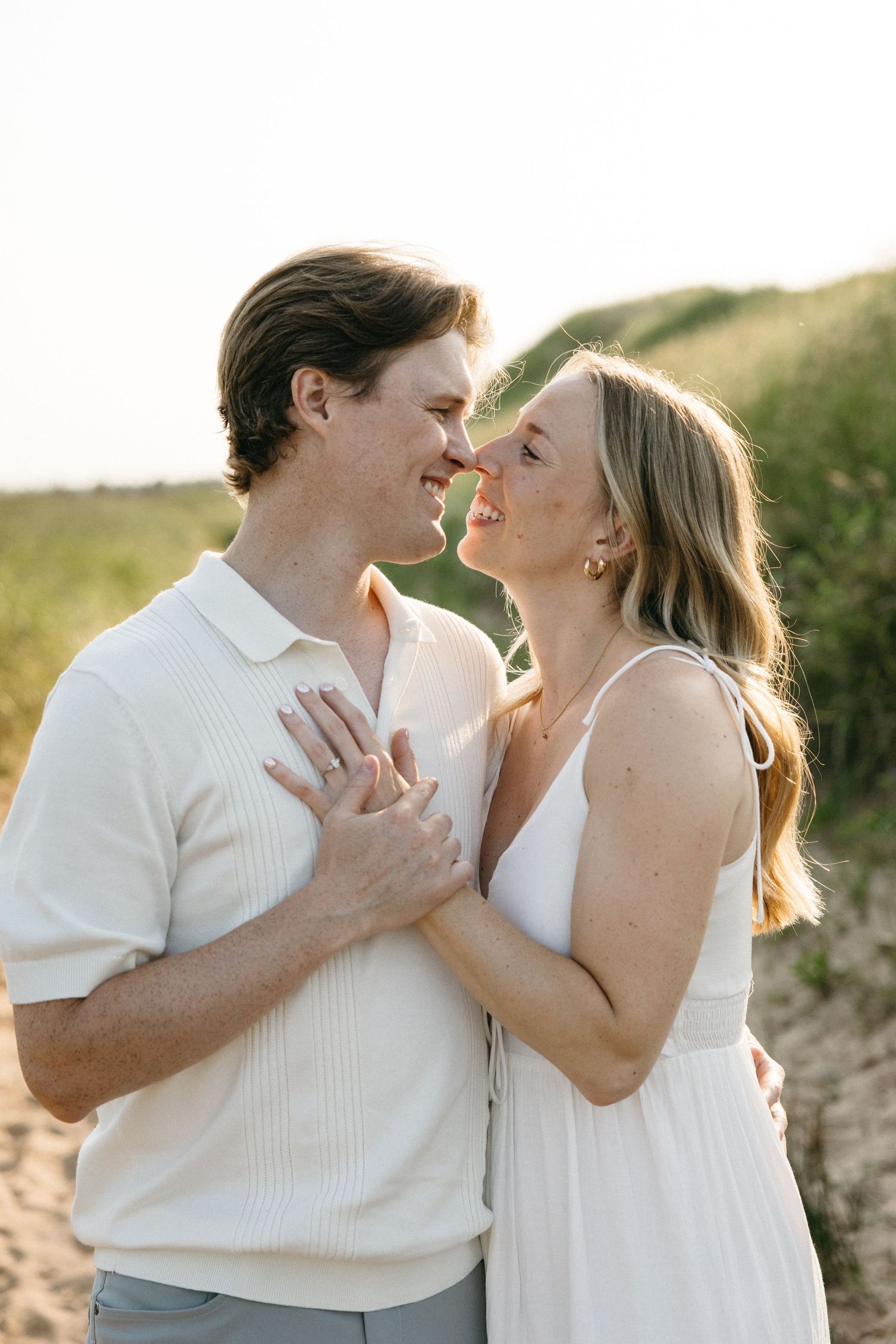 Smiling couple almost kissing on sandy beach path at golden hour, woman in white dress, man in white polo.
