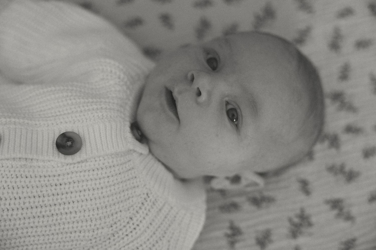Black and white portrait of an infant wearing a white knit sweater against floral bedding.