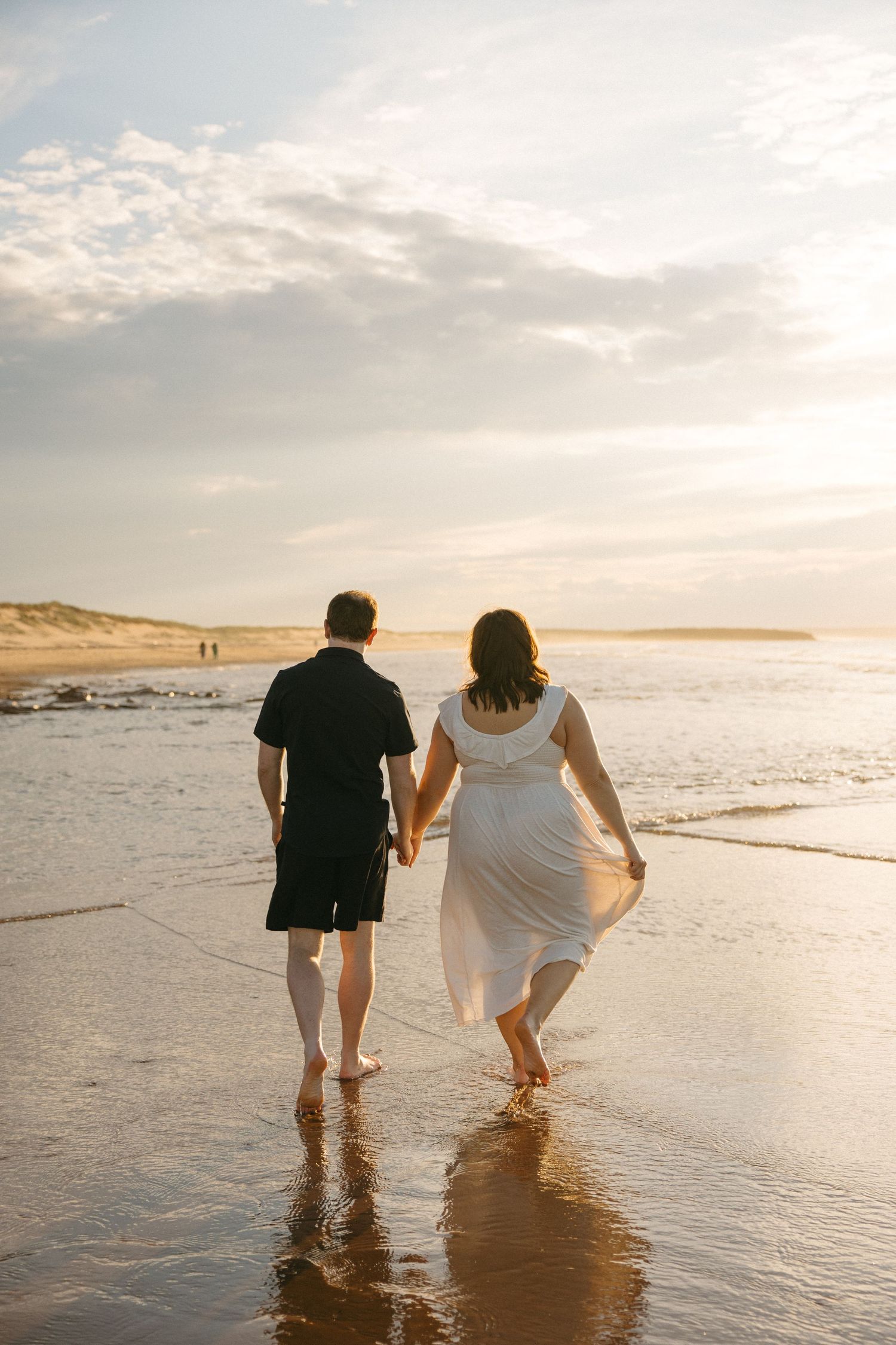 Couple holding hands walking barefoot on beach at sunset, woman in white dress.