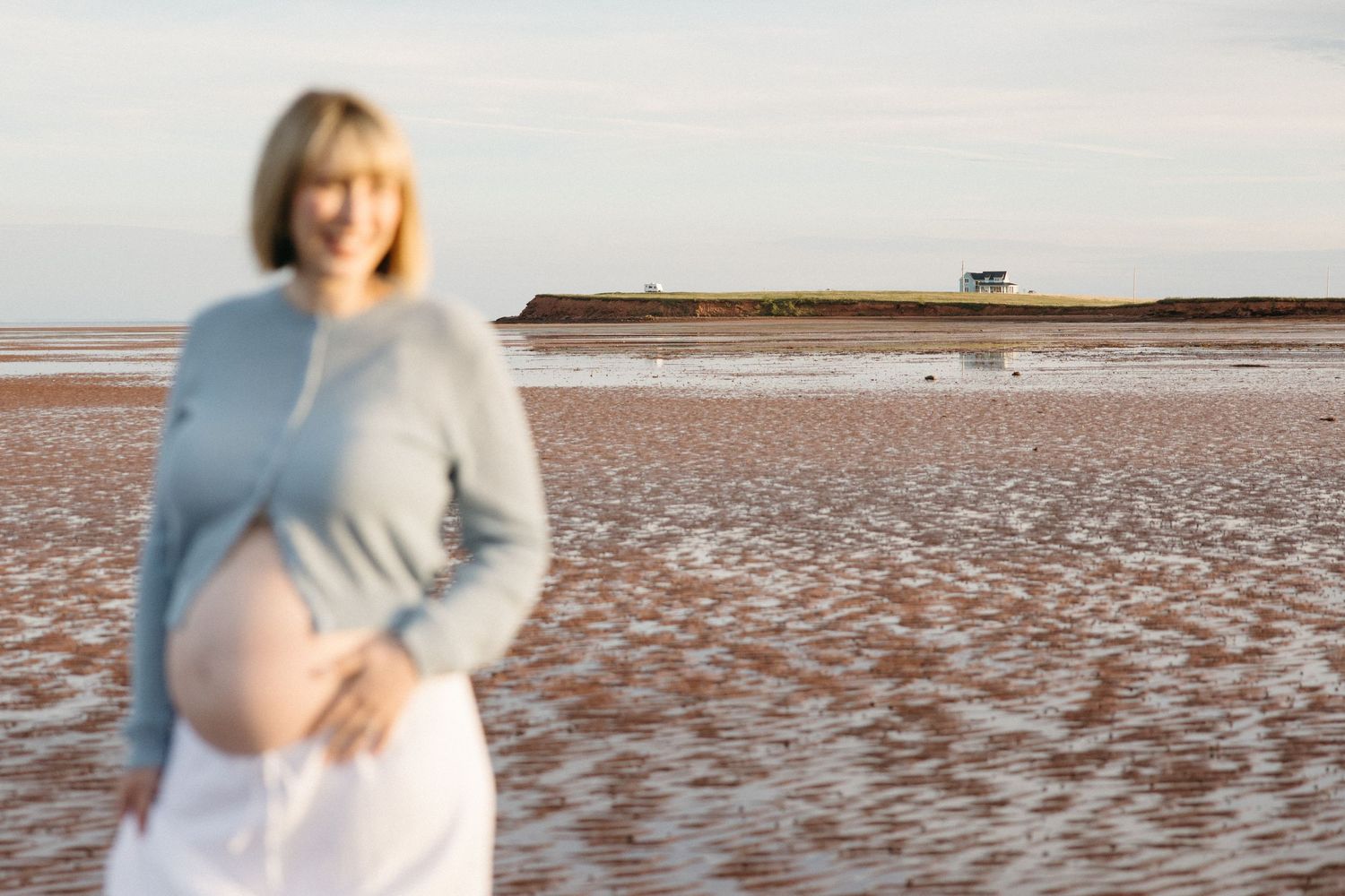 Smiling pregnant woman in blue sweater cradling baby bump on a sandy beach at golden hour.