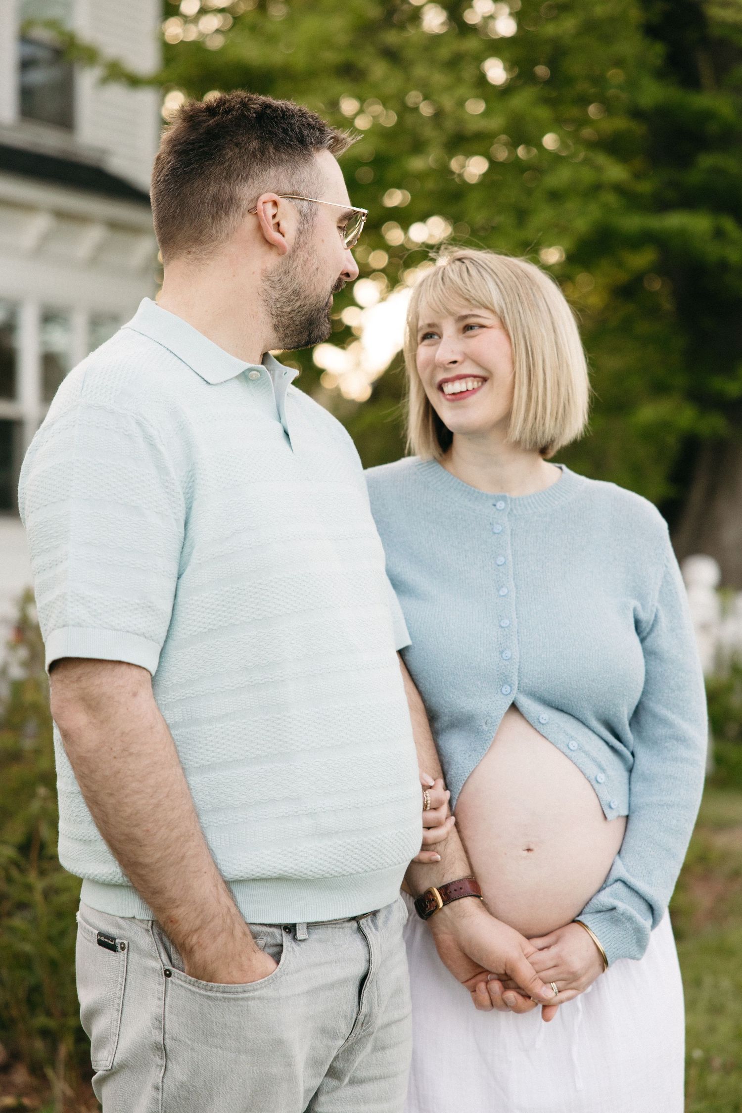 Smiling pregnant woman in blue cardigan holds baby bump while partner looks at her lovingly outdoors.