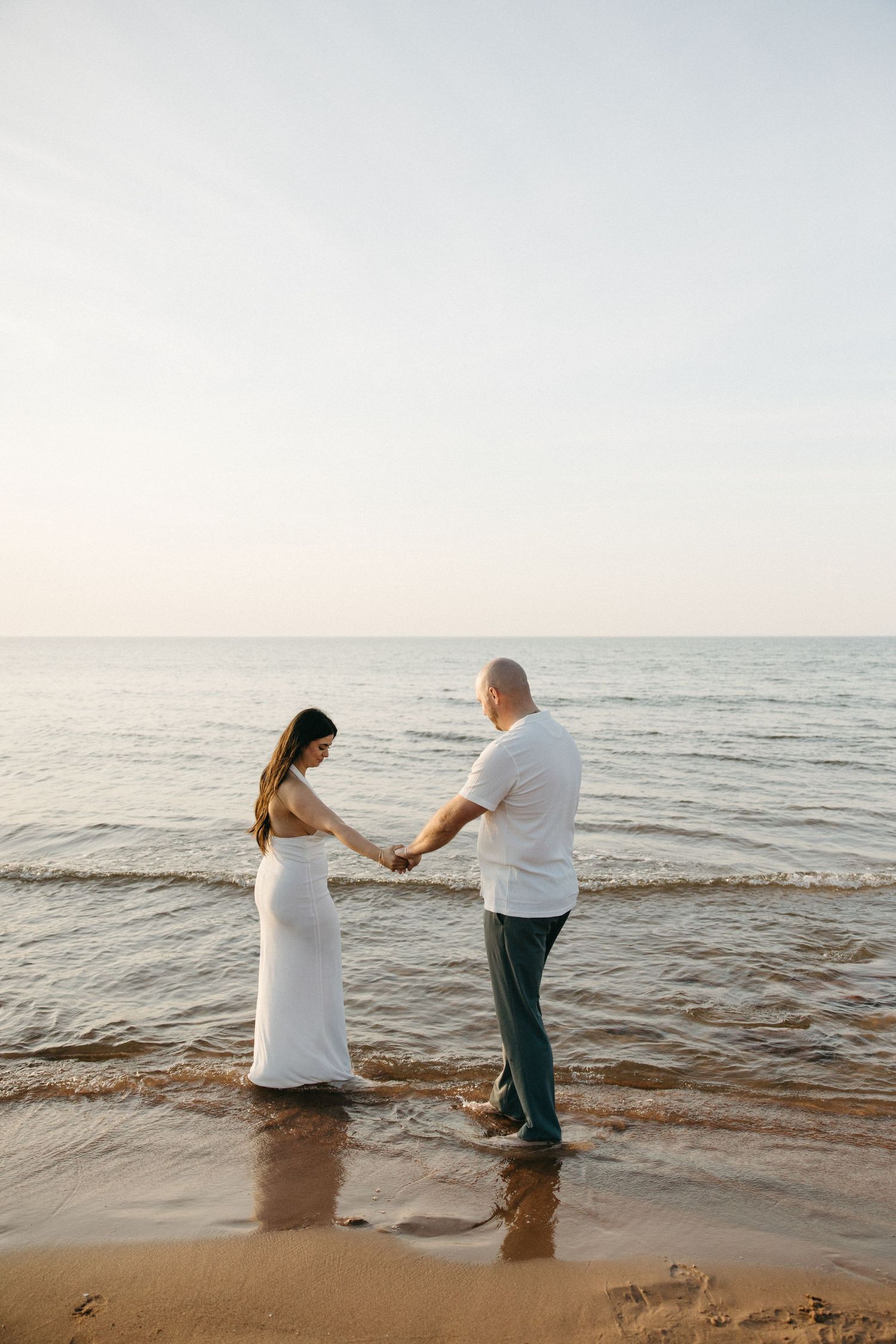 Couple holding hands while standing in shallow ocean water at sunset, woman in white dress, man in jeans.