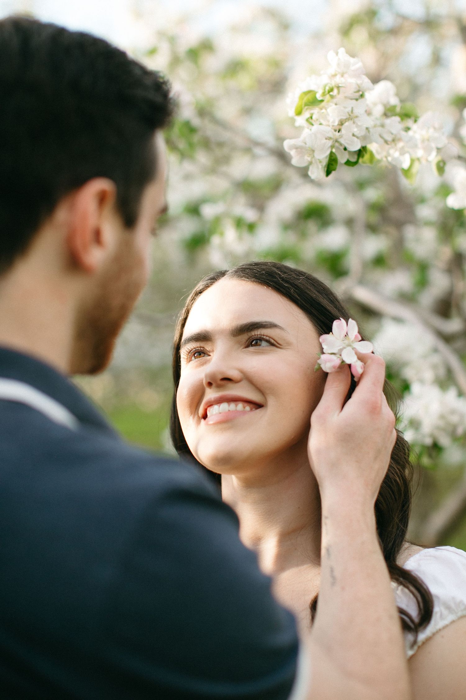 A man places a white flower in a smiling woman's hair near blooming apple trees.