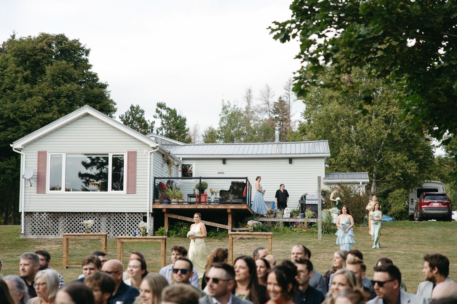 Wedding guests gather outdoors on a lawn surrounded by trees for an intimate ceremony.