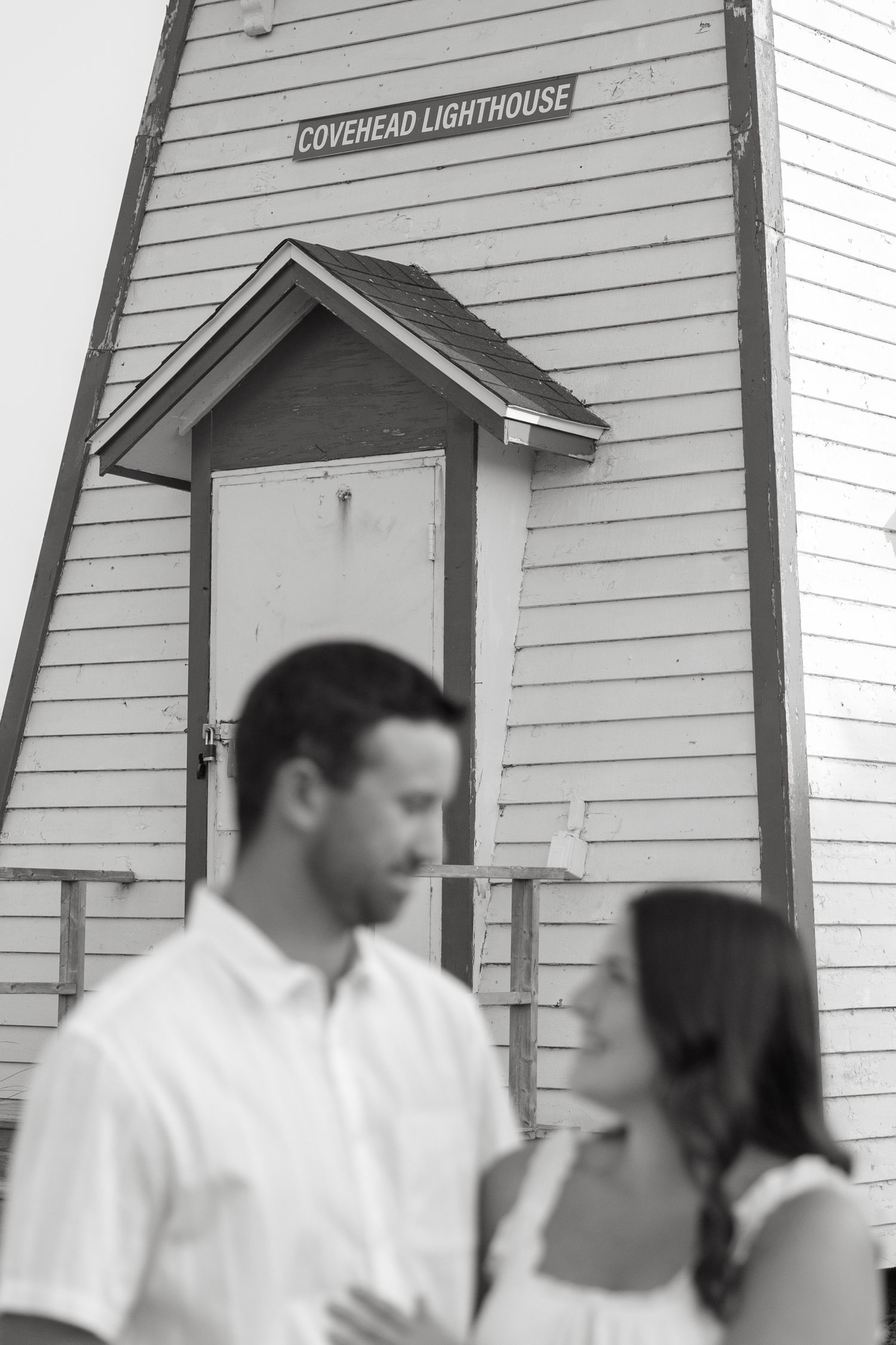A couple poses in front of the Covehead Lighthouse sign in black and white.