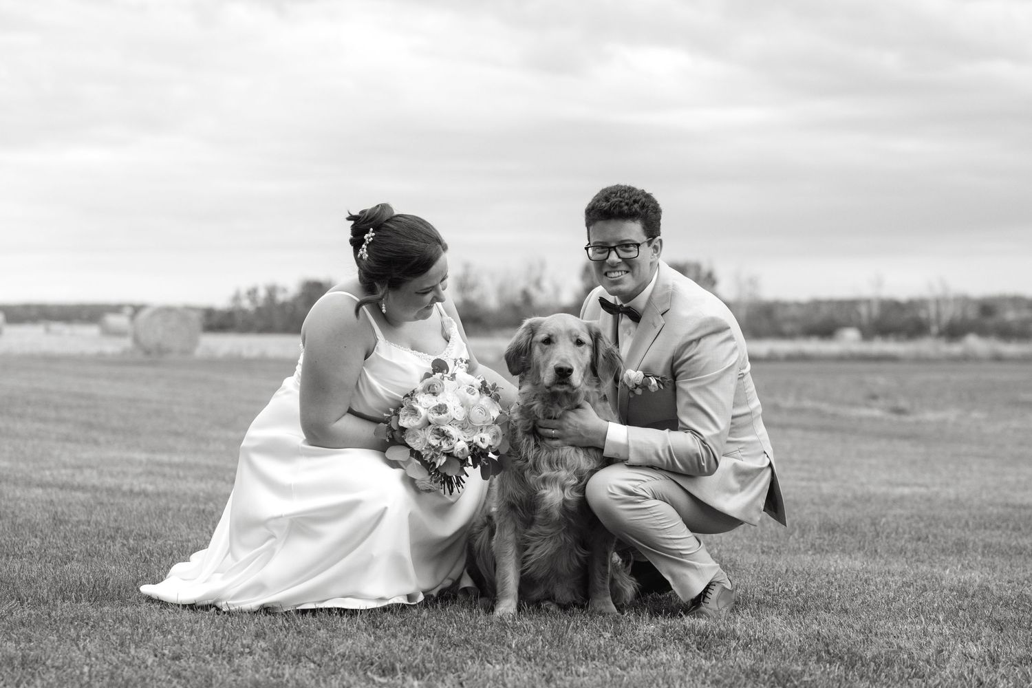 Black and white photo of newlywed couple posing with their golden retriever dog on grass at wedding.