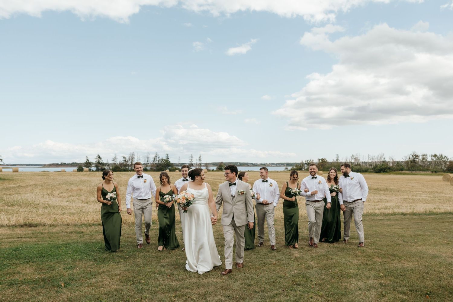 Wedding party in green dresses and gray suits posing in an open field with water views behind them.