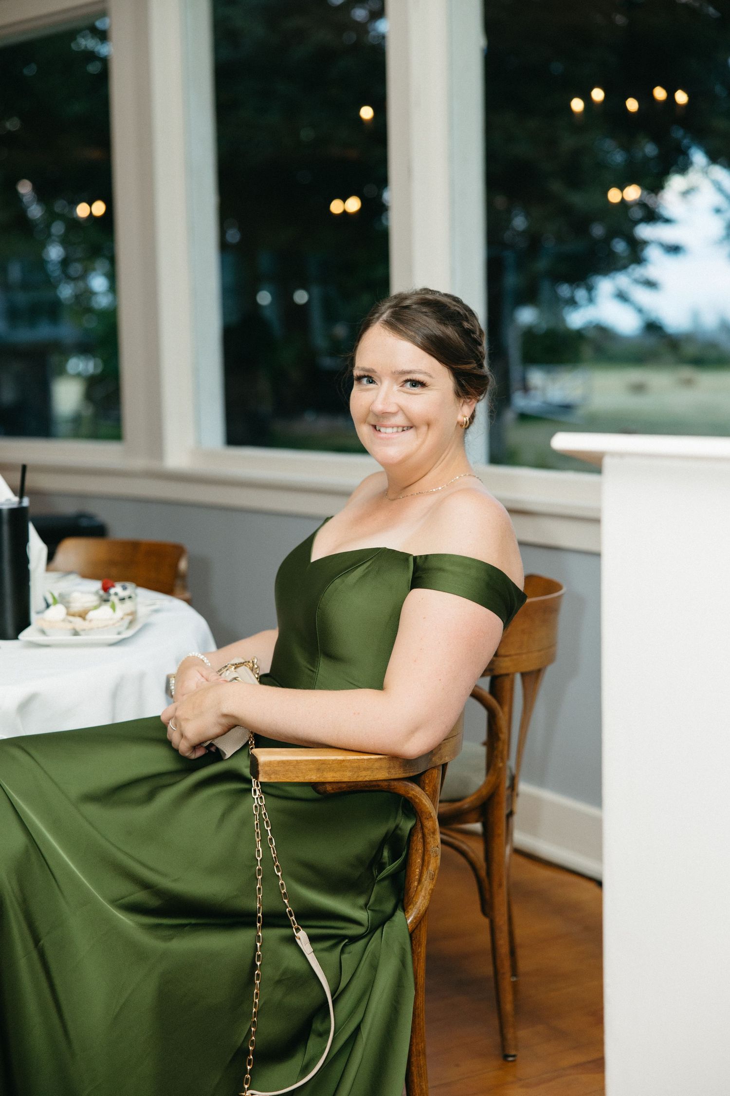 Bridesmaid in elegant off-shoulder olive green gown smiles while seated at a reception table.