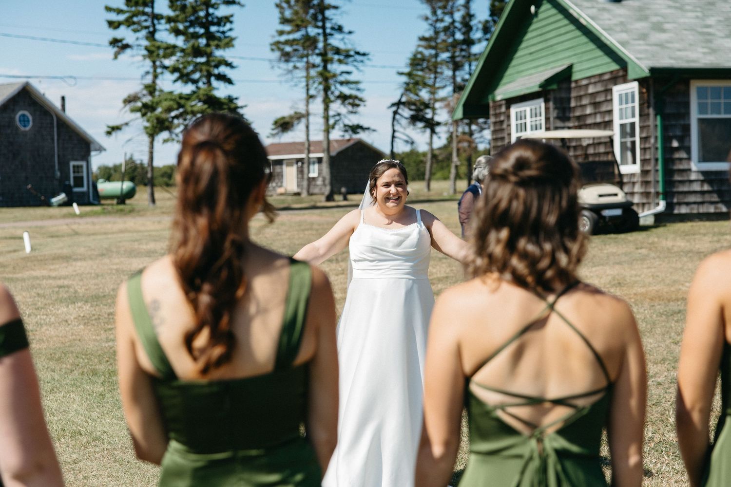 A bride in a white gown faces her bridesmaids in olive green dresses on a sunny lawn.