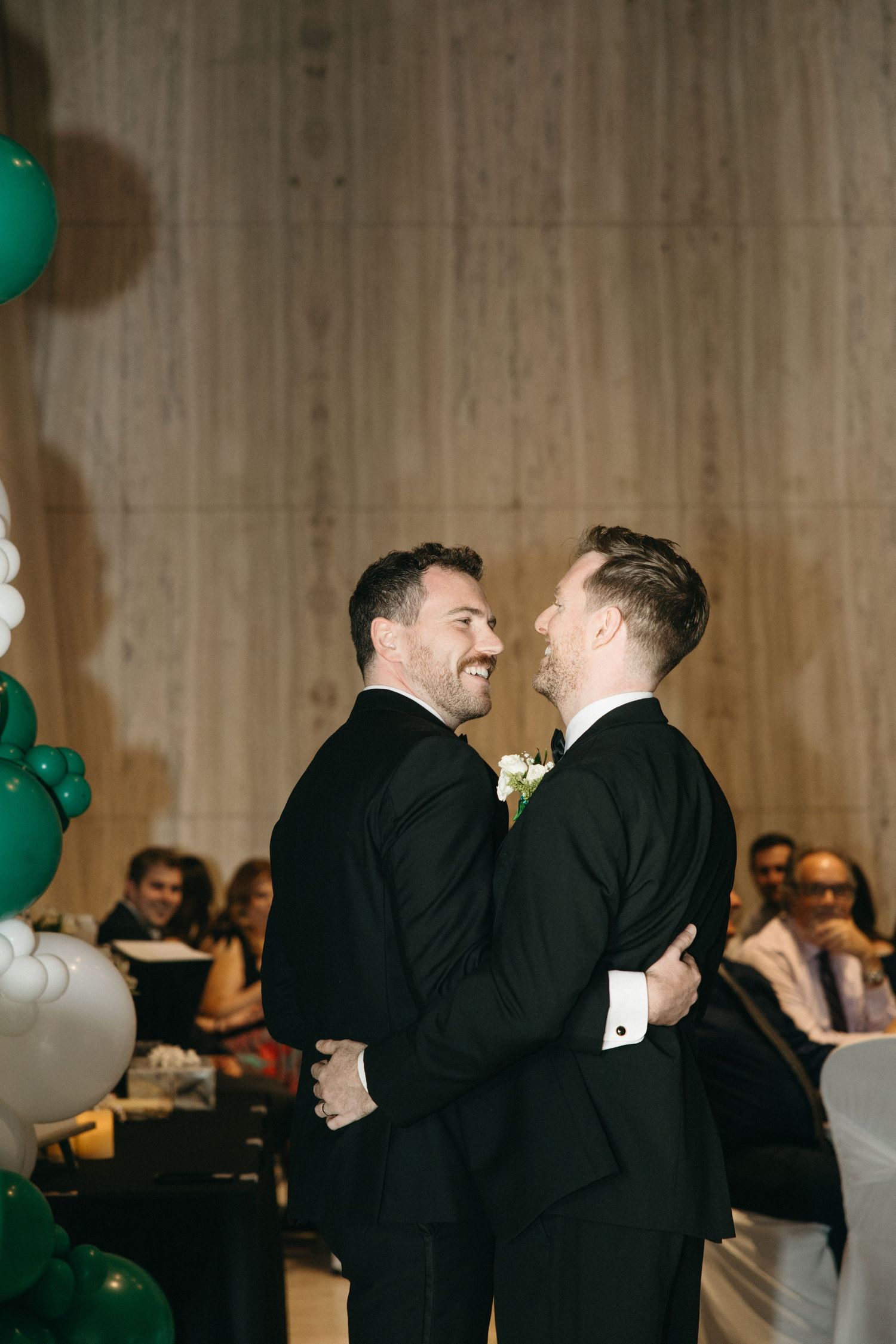 Two people in black suits sharing a joyful moment at a wedding with green and white balloons.