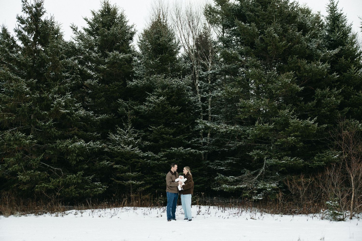 A couple stands together holding their baby in a snowy forest with tall evergreen trees.